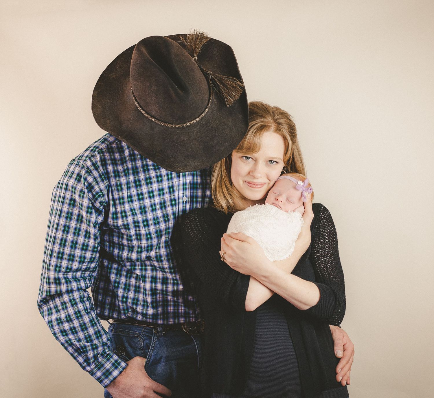 Intimate family portrait with plaid shirt, cowboy hat, and tender embrace against neutral background.