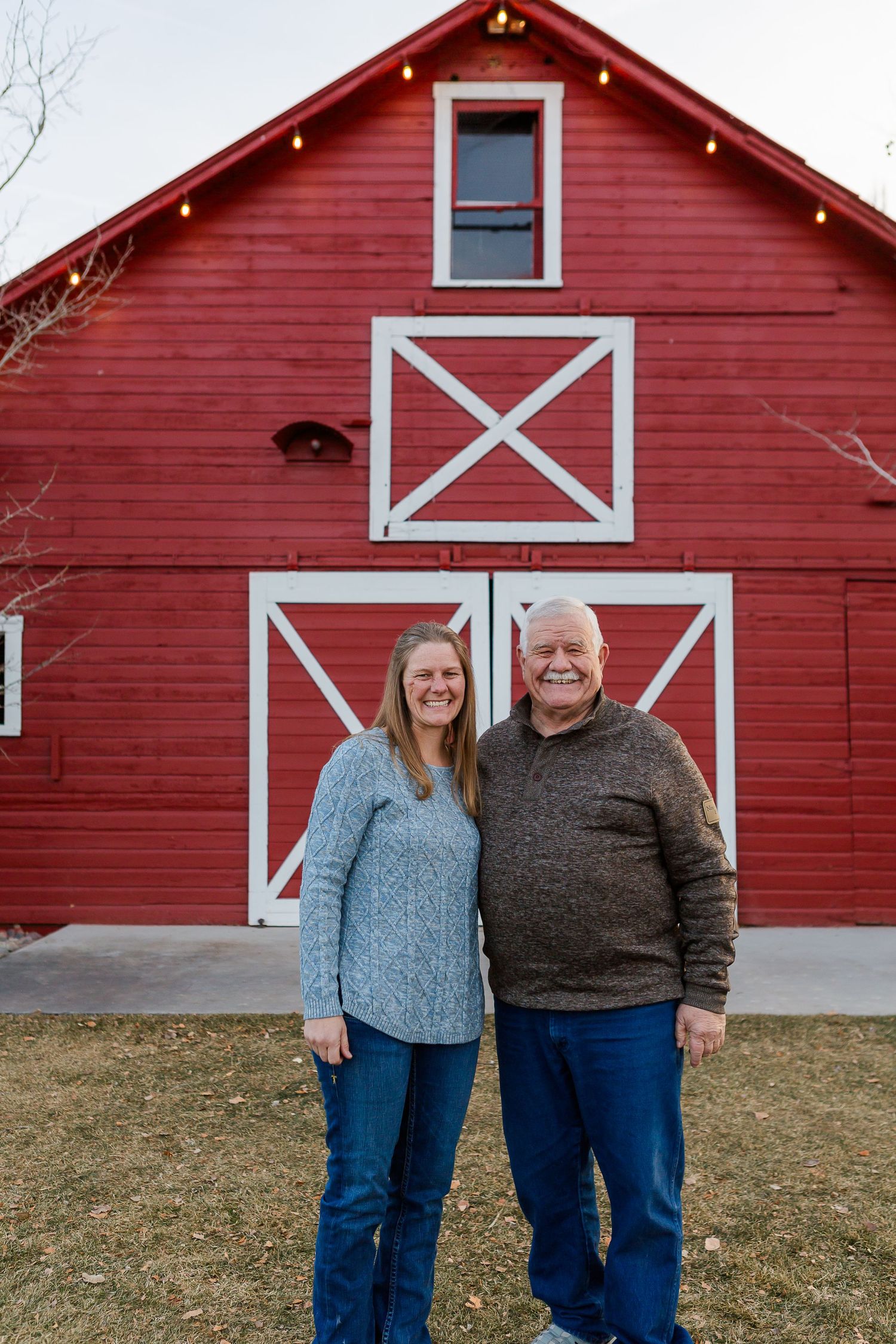 Two people standing side by side in front of a red barn with white X-pattern doors on a winter day.