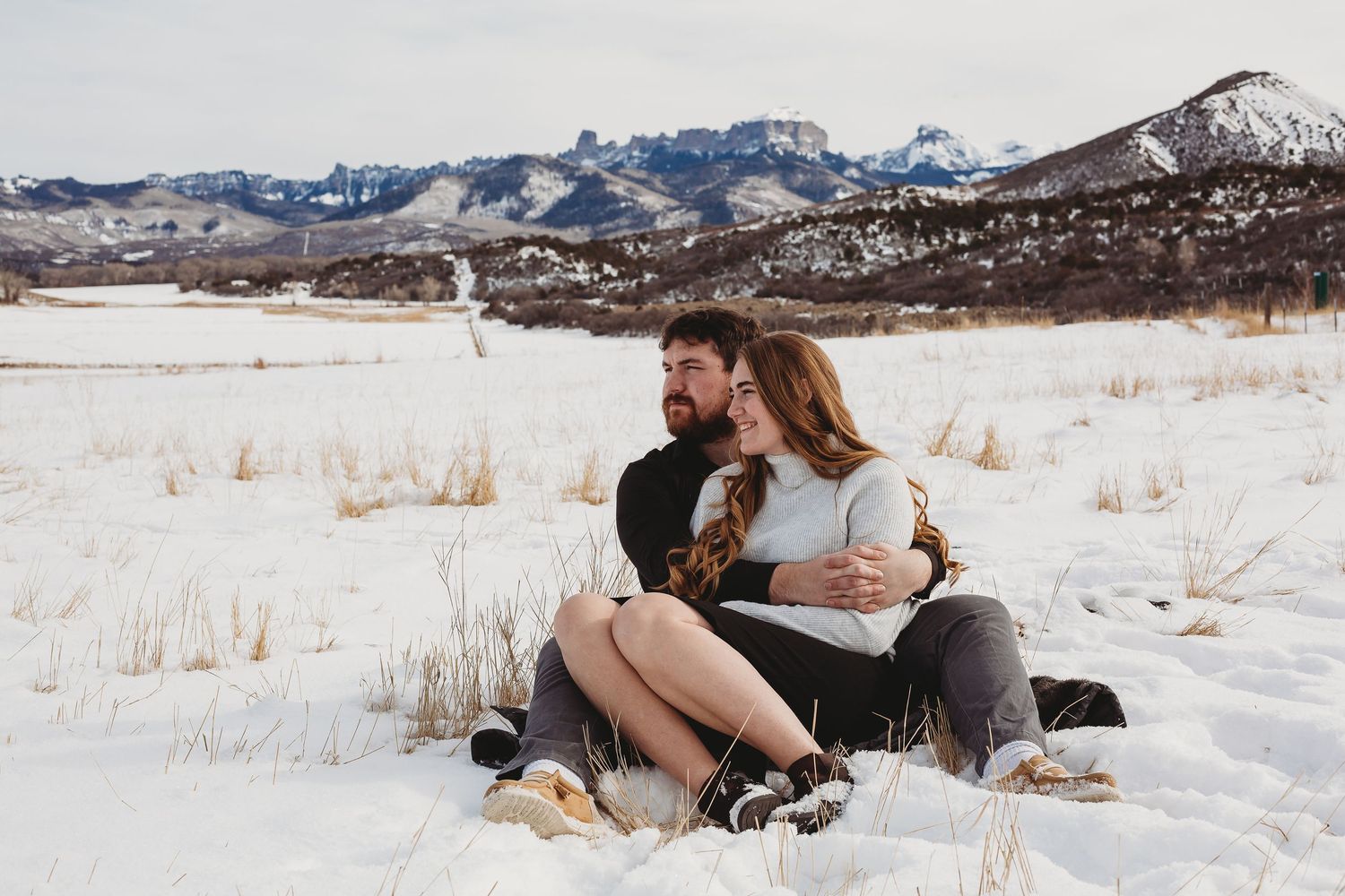 Couple sitting together on snowy ground with mountain vista in background.