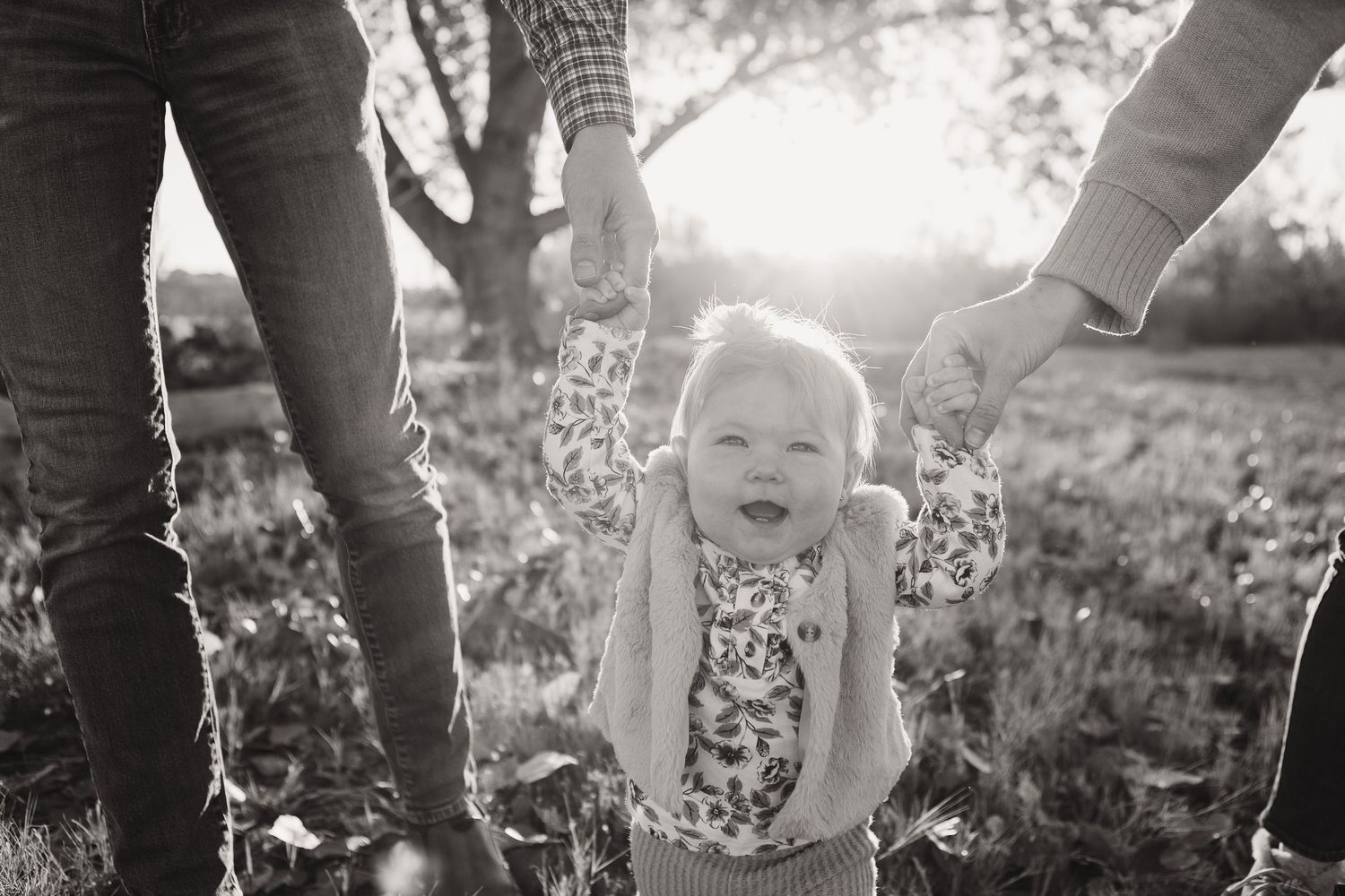 A toddler holds hands with parents in a sunlit field during a black and white photo session.