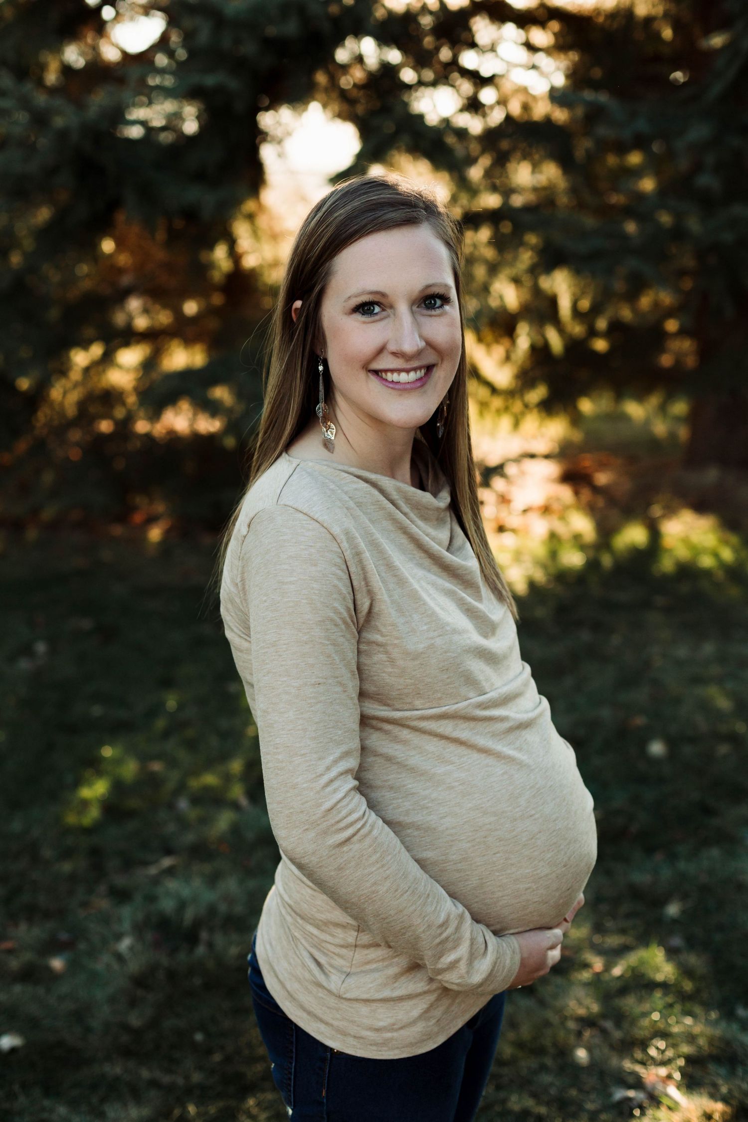 Maternity portrait of an expectant mother in a beige sweater against a sunlit evergreen forest backdrop.