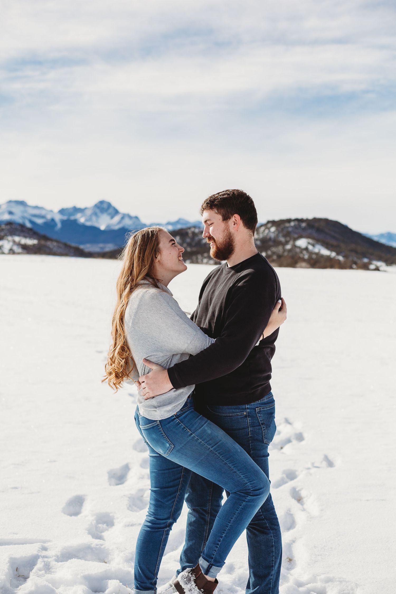A couple shares a romantic embrace while standing in deep snow with mountains behind them.
