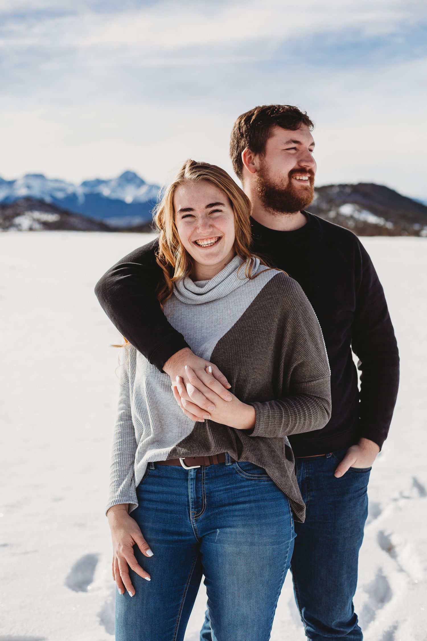 A joyful couple embraces while standing in a snowy field with mountains in the distance.