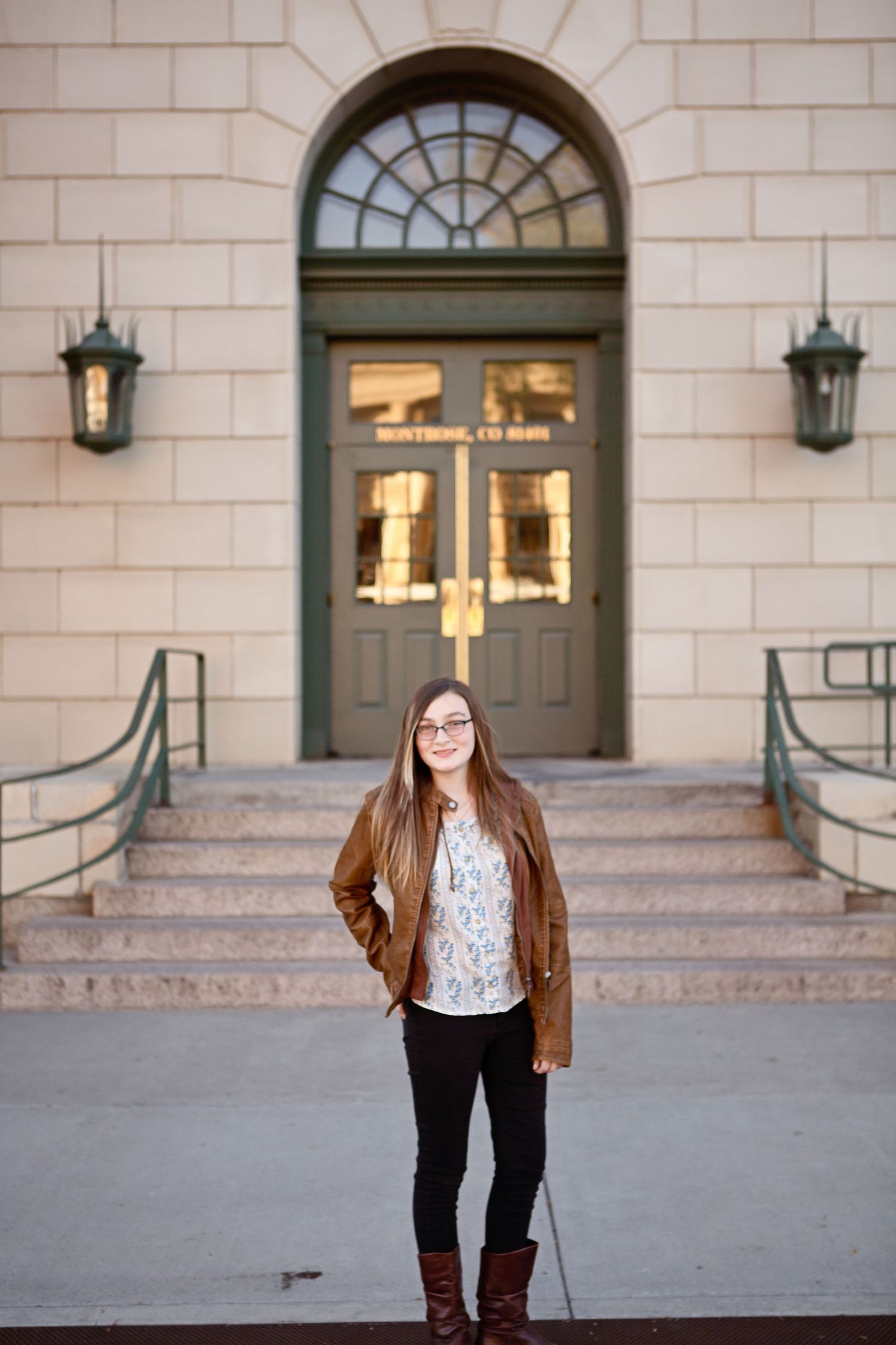 A person in a brown jacket stands in front of an elegant building entrance with arched doorway.