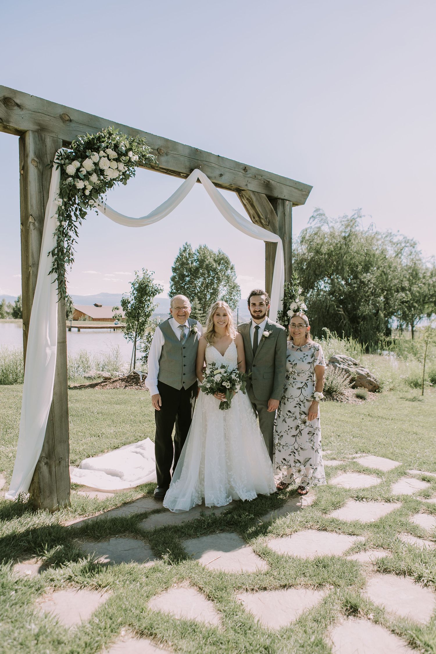 Wedding party poses under wooden arch decorated with white fabric and greenery in outdoor ceremony space.