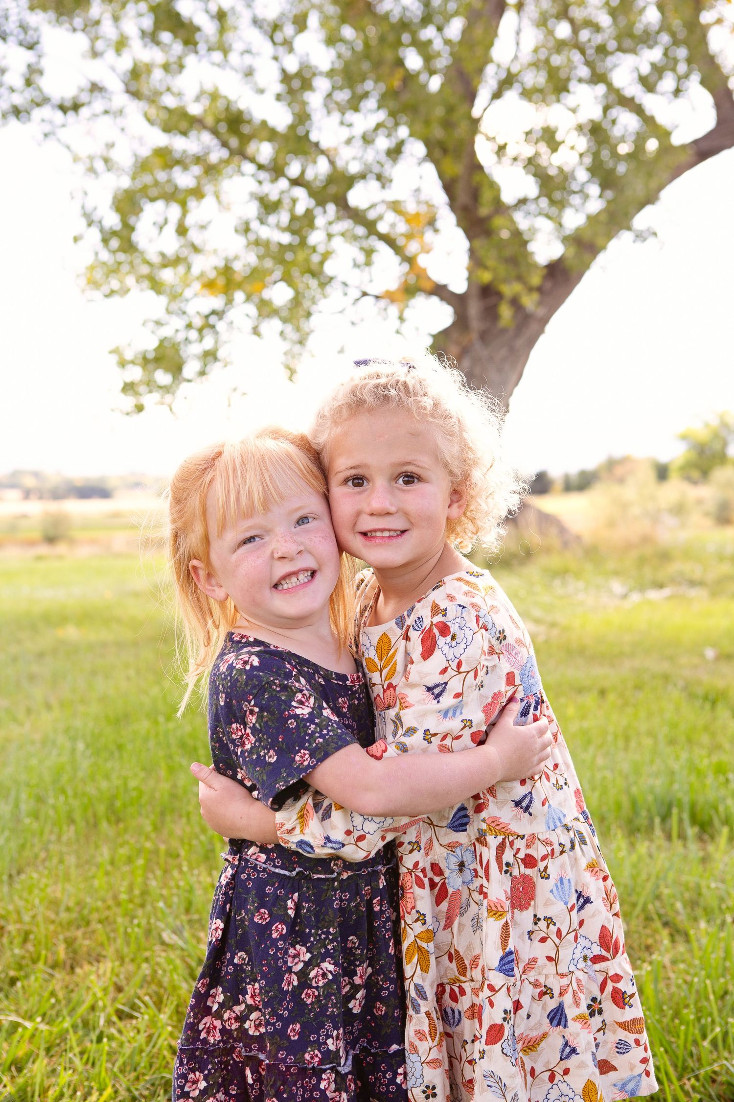 Two children share a joyful hug while wearing floral dresses in a sunlit field.