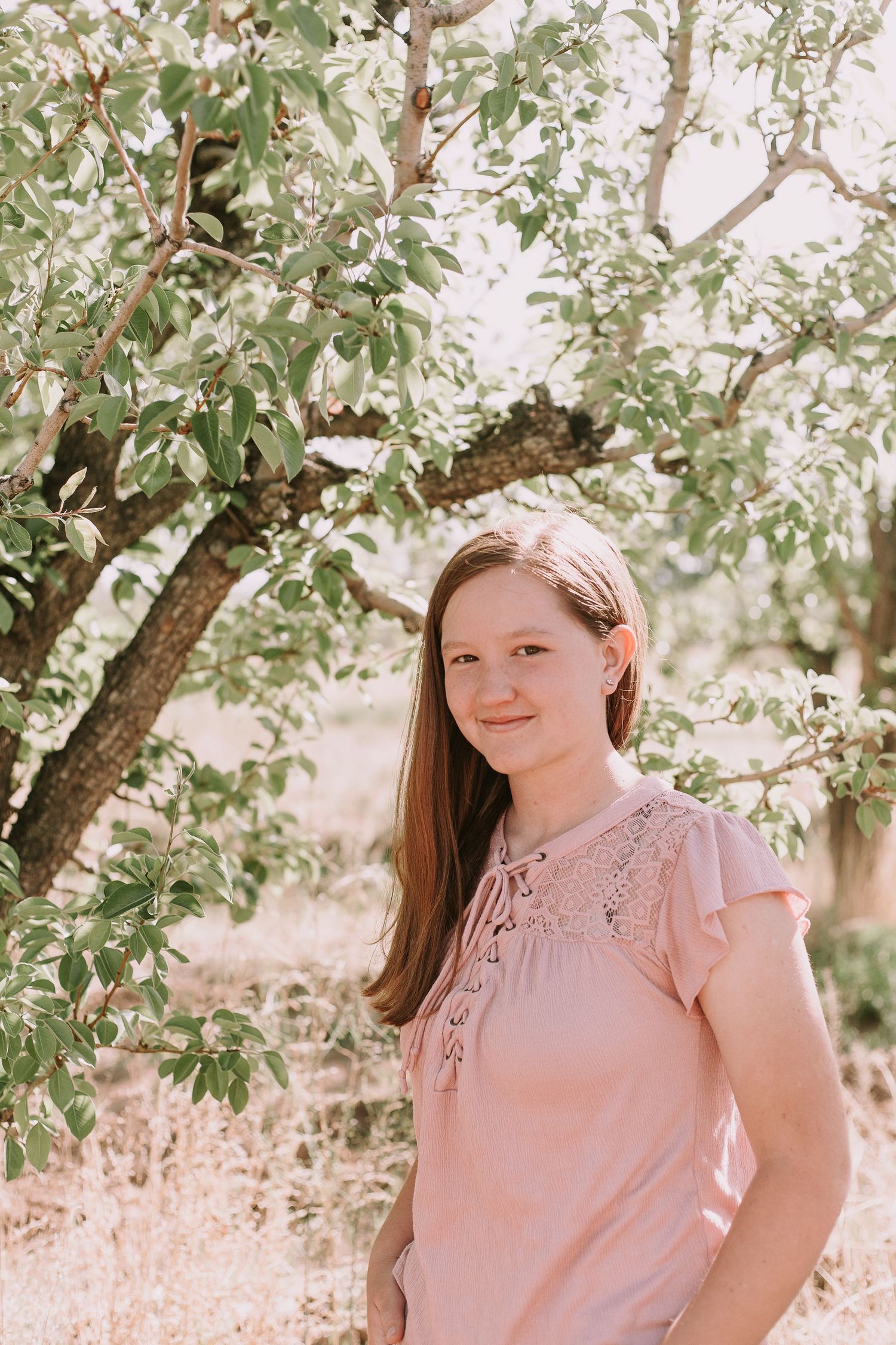 A person in a pink lace blouse poses against a leafy tree backdrop in soft natural lighting.