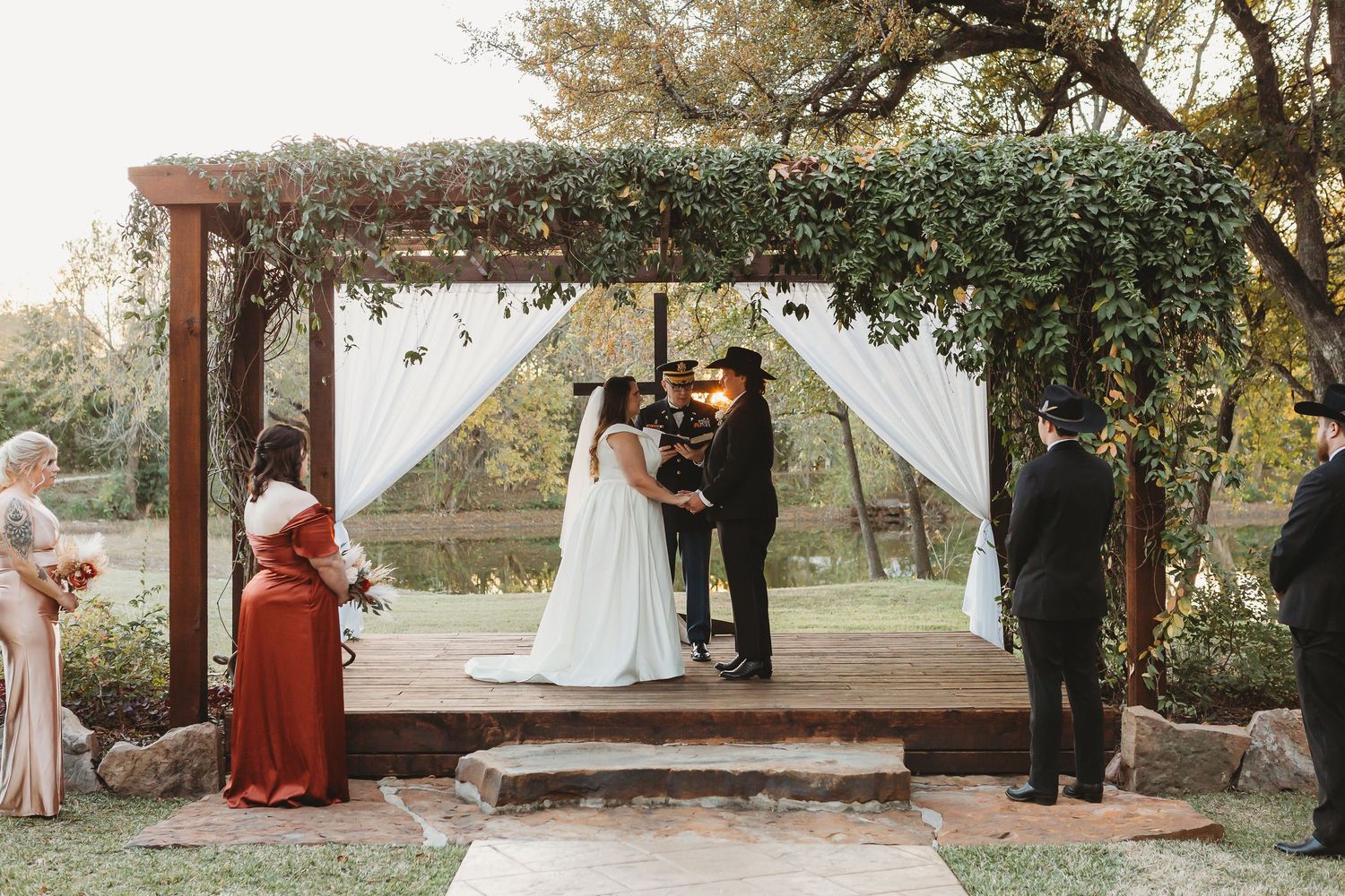 Wedding ceremony takes place under rustic wooden arch decorated with greenery and white fabric.