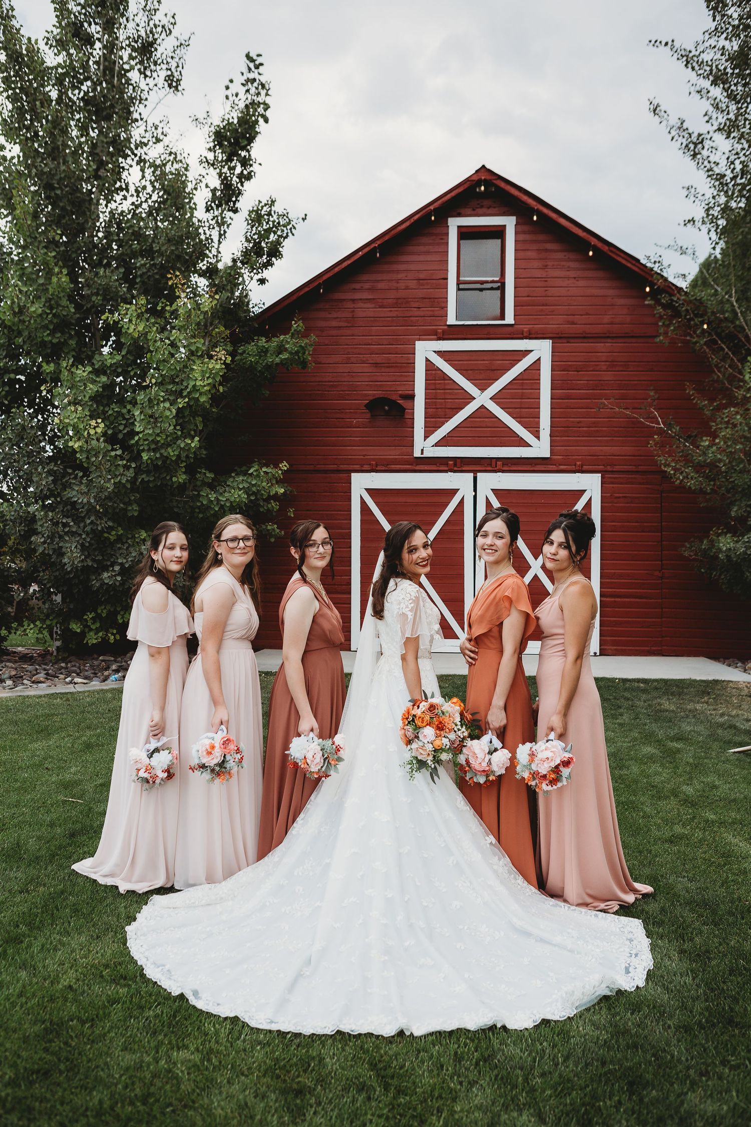 Wedding party poses in front of a red barn with white trim wearing earth-tone dresses.