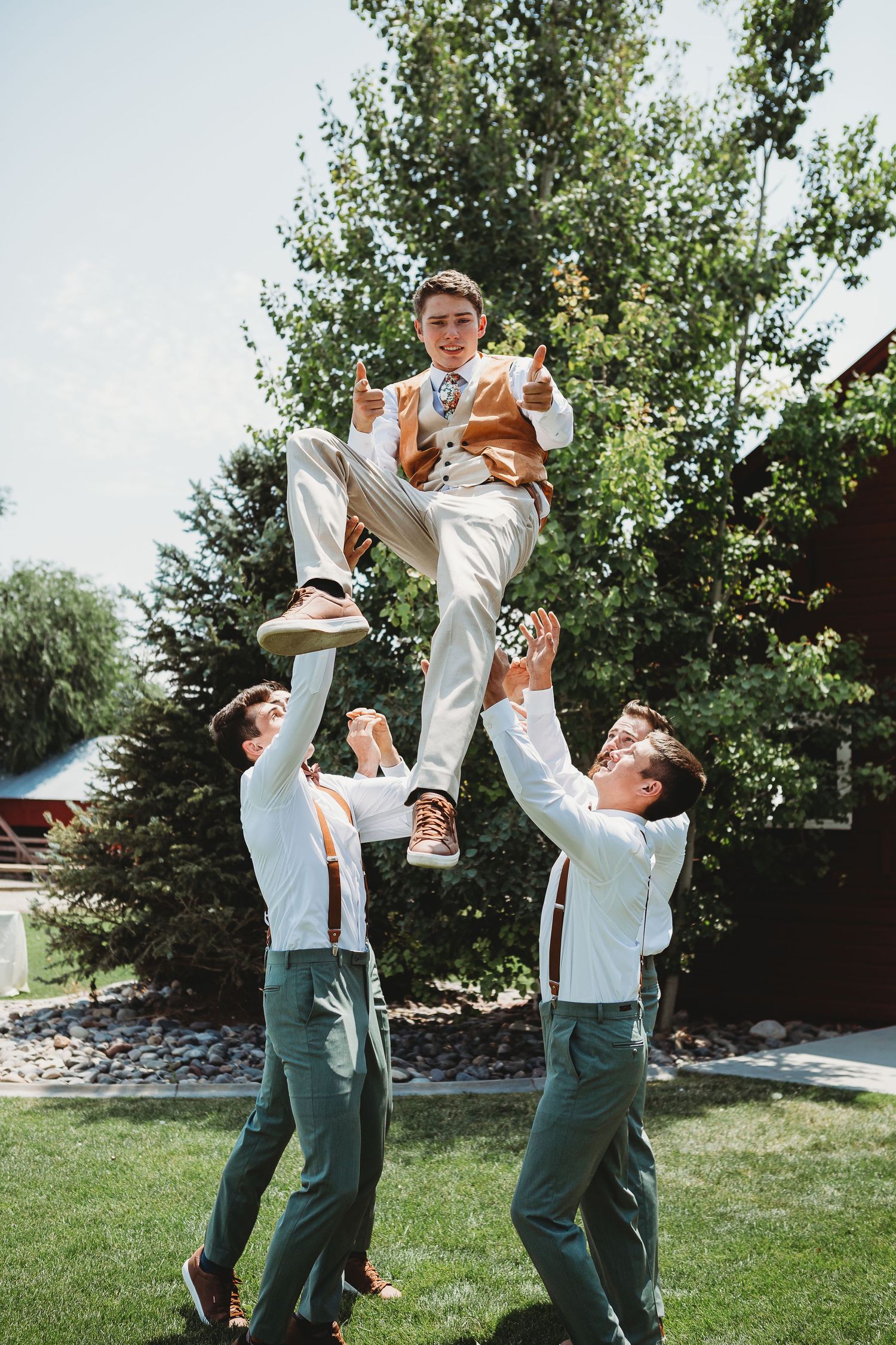 Groomsmen playfully lift another member of the wedding party in front of trees.