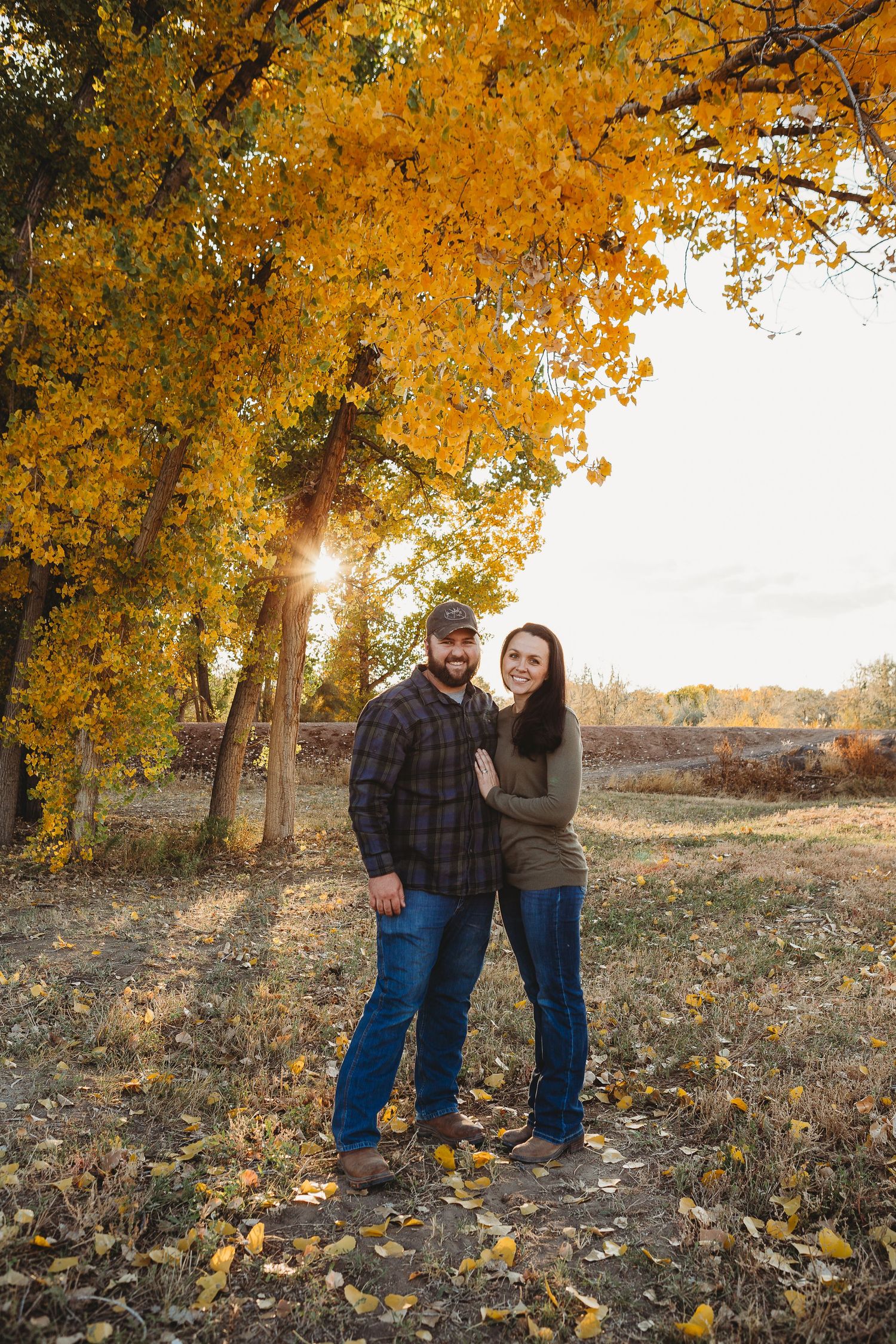 A couple stands beneath vibrant yellow autumn trees as sunlight filters through the leaves.