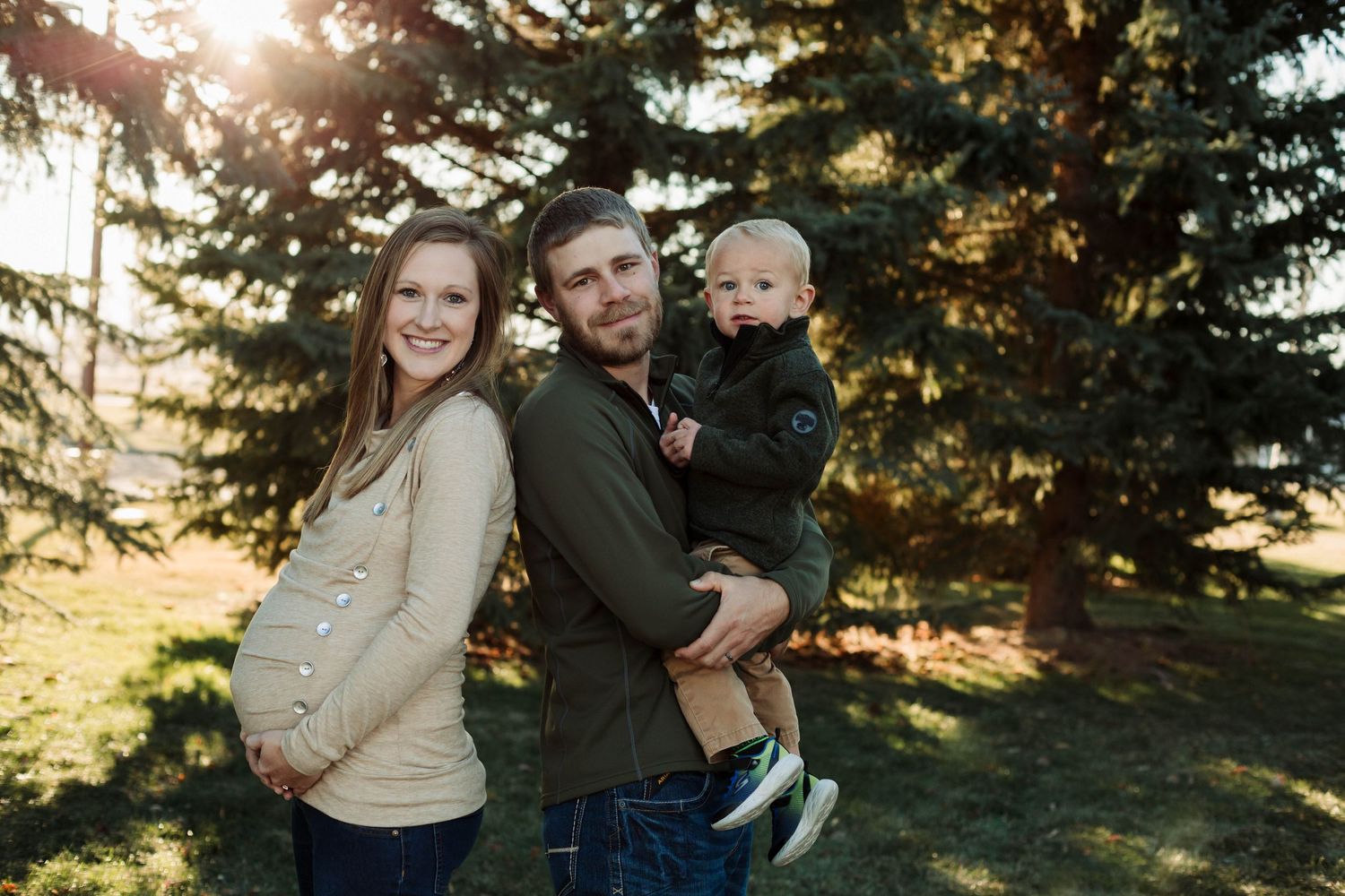 Family of three poses outdoors with pregnant mother in cream sweater and father holding toddler.