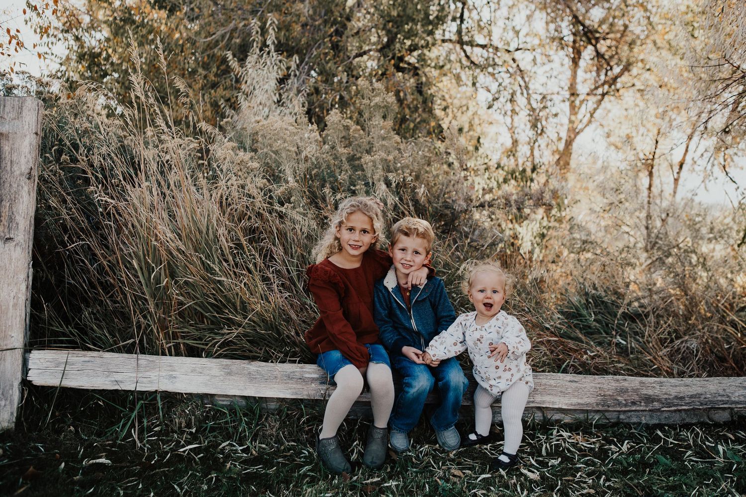 Three children sit together on a rustic wooden bench against a natural outdoor backdrop at sunset.