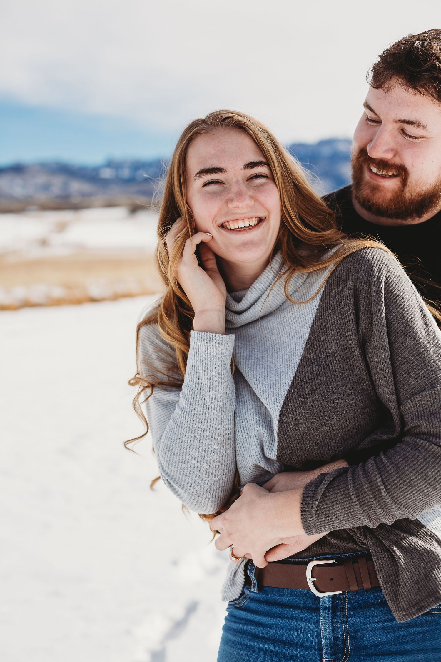 A joyful couple embraces while posing in a winter mountain landscape.
