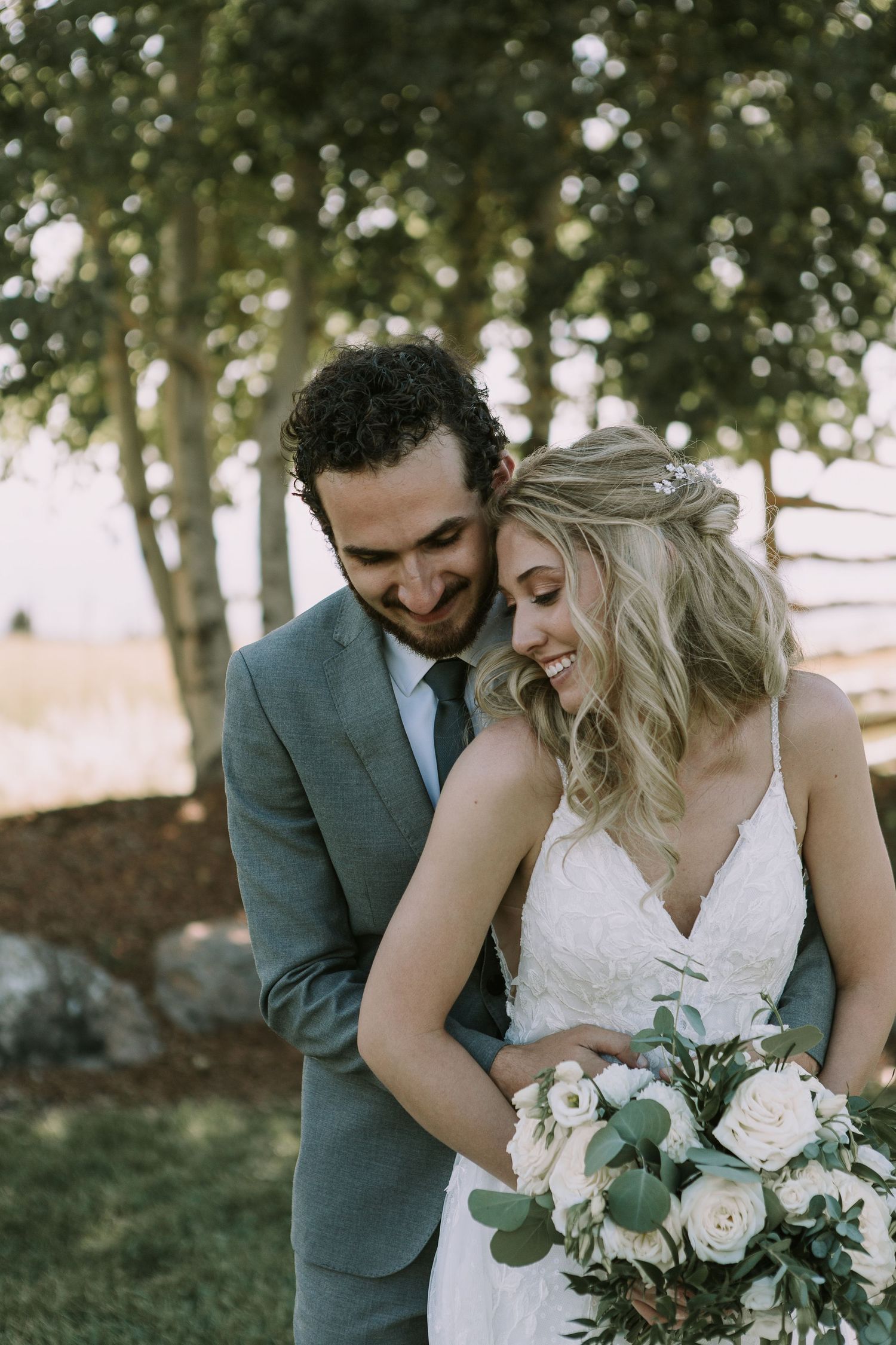 Intimate wedding moment captured under trees with the bride holding a delicate rose bouquet.