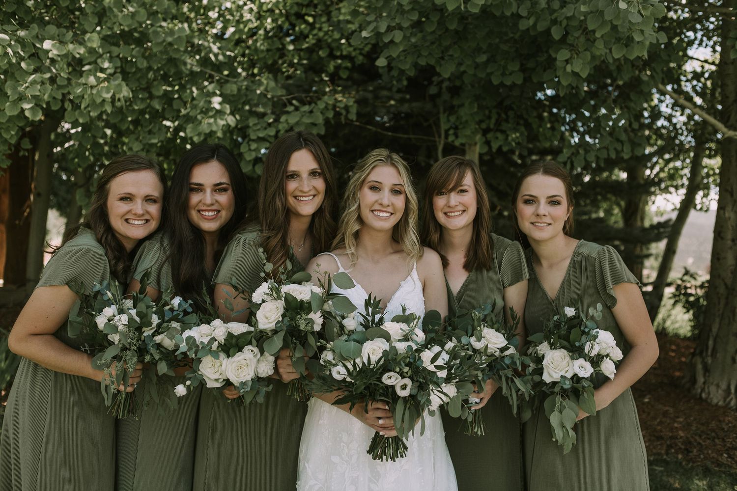 Bride poses with bridesmaids in olive green dresses holding white floral bouquets.