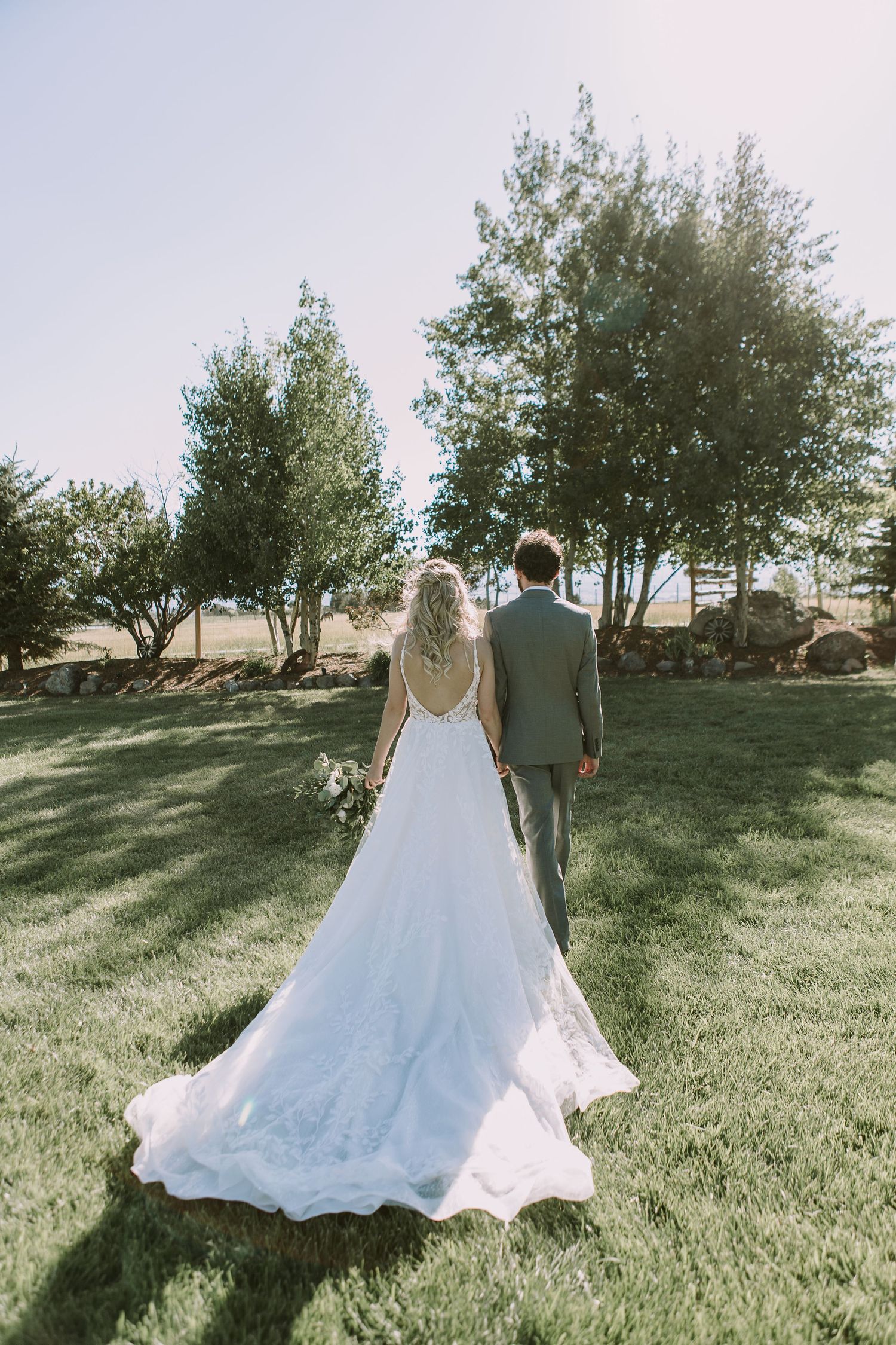 Bride in flowing white dress walks with groom across a sunlit lawn with trees in background.