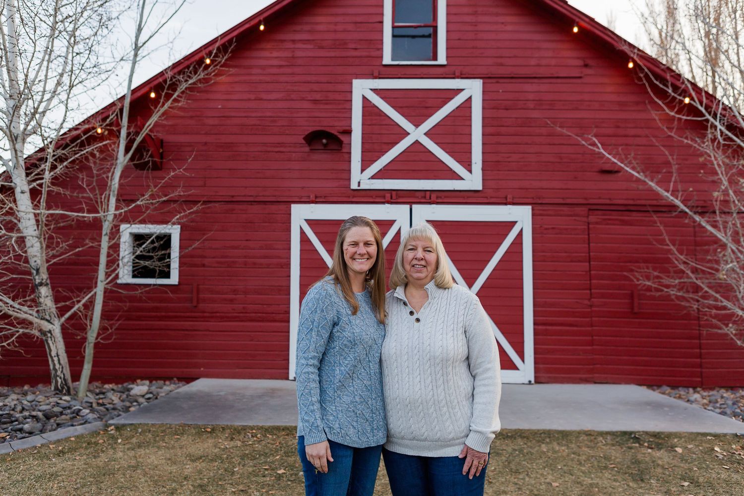 Two people standing together and smiling in front of a red barn with white cross pattern decorations.