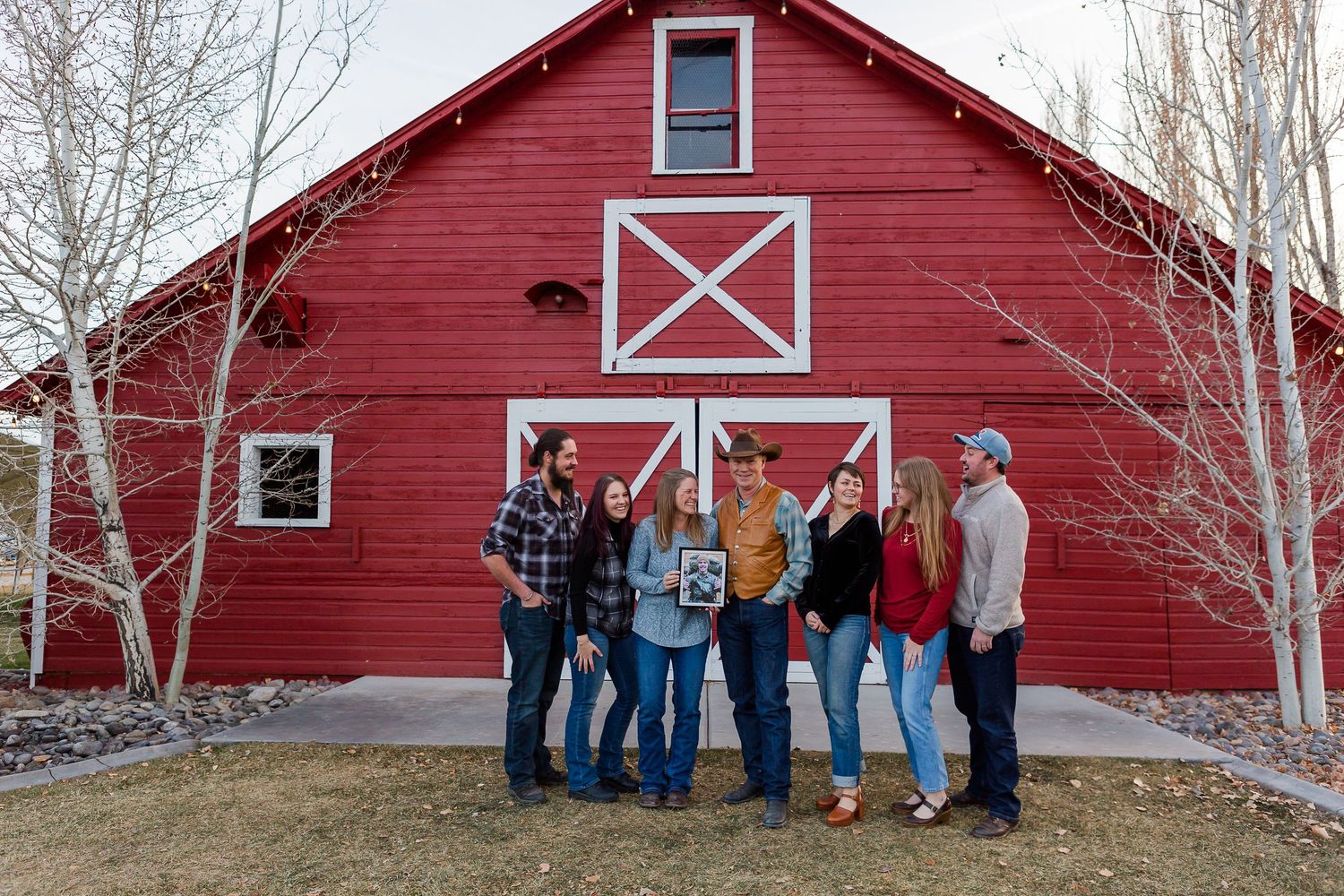 Family gathering posed casually in front of bright red barn with white decorative elements.