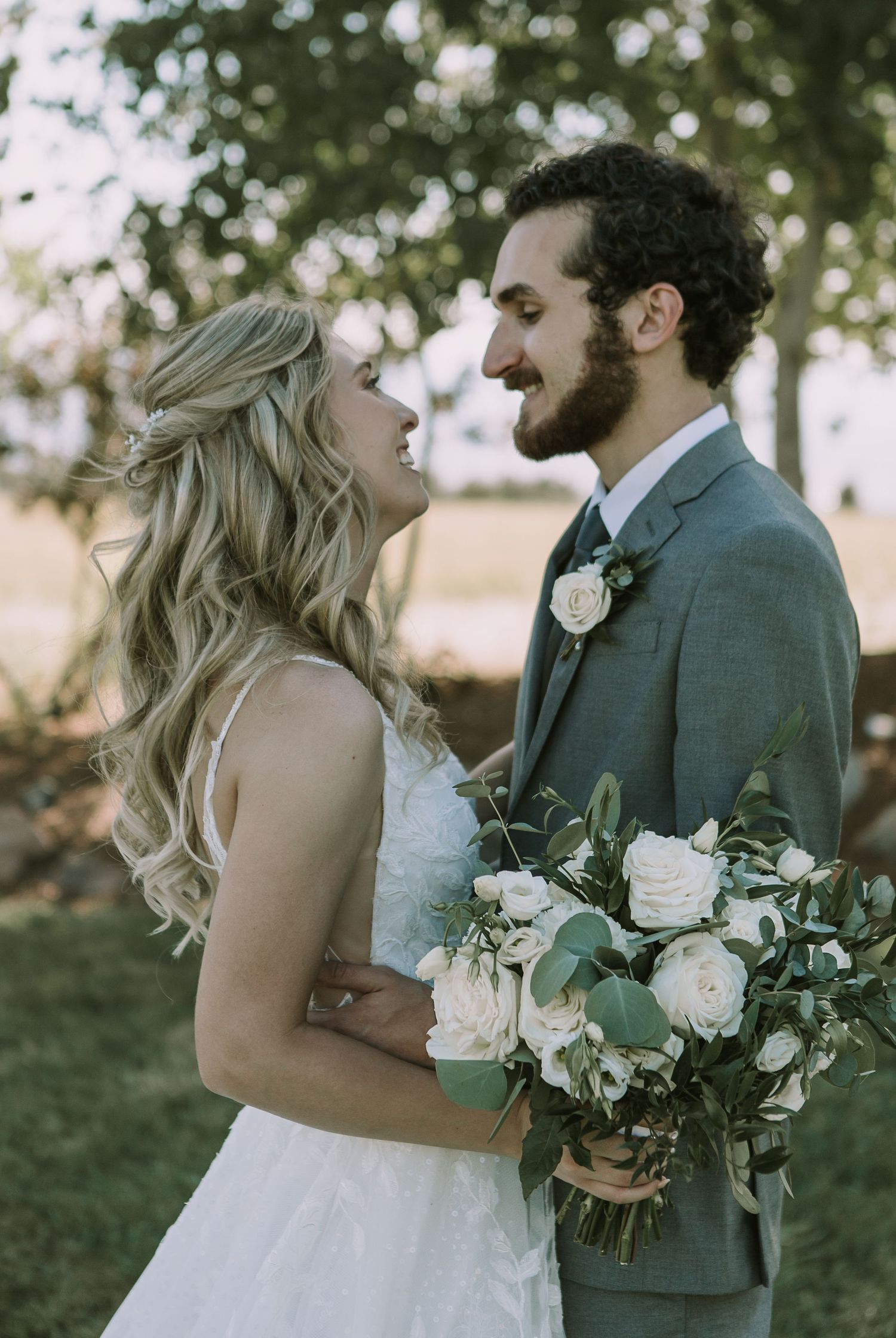 Bride and groom share joyful laughter while holding a white rose bouquet against a tree-lined backdrop.