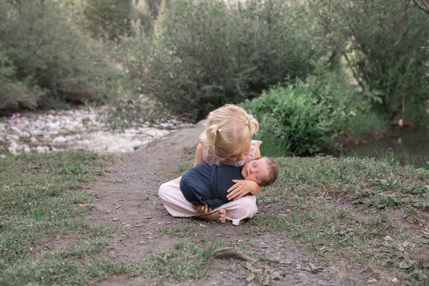A tender outdoor family moment captured on a dirt path beside a stream in a natural forest setting.