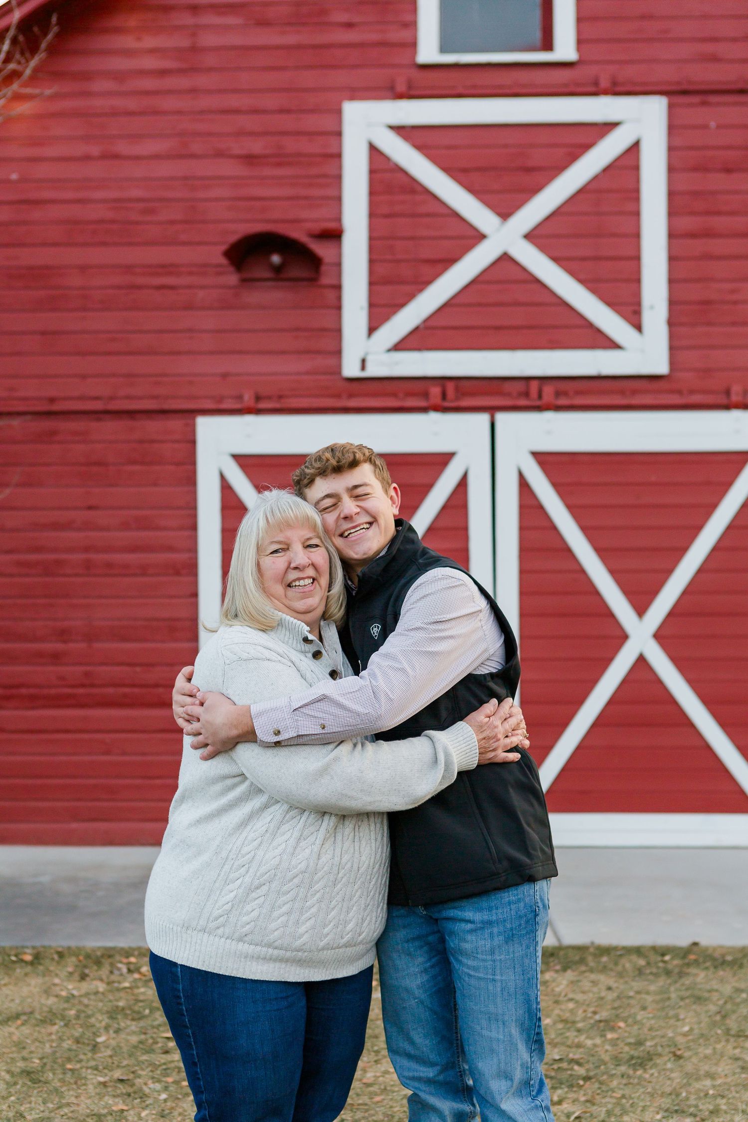 Two people share a happy hug while posing in front of a red barn with white trim.