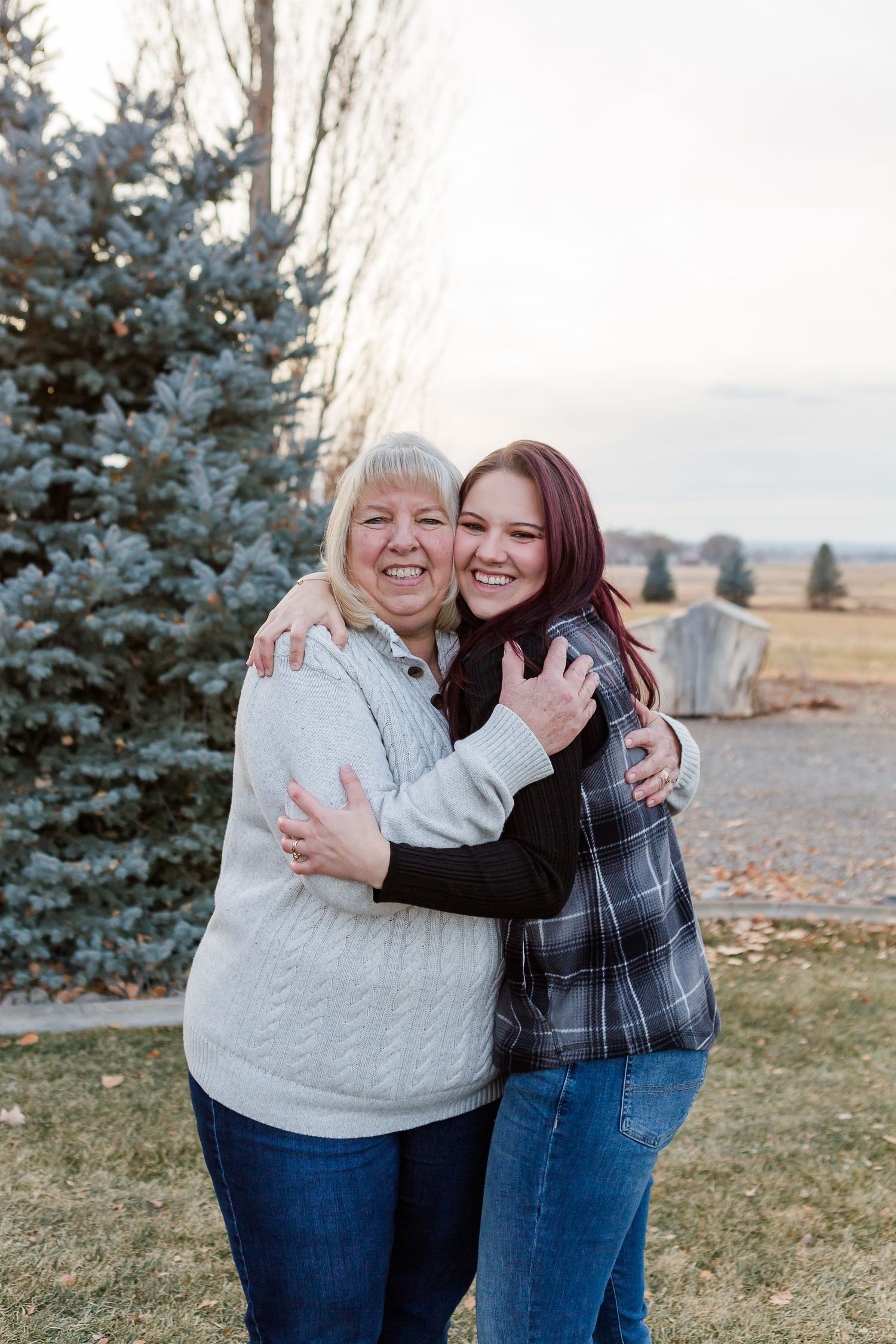 Two people embrace and smile while standing in front of an evergreen tree on a rural property.