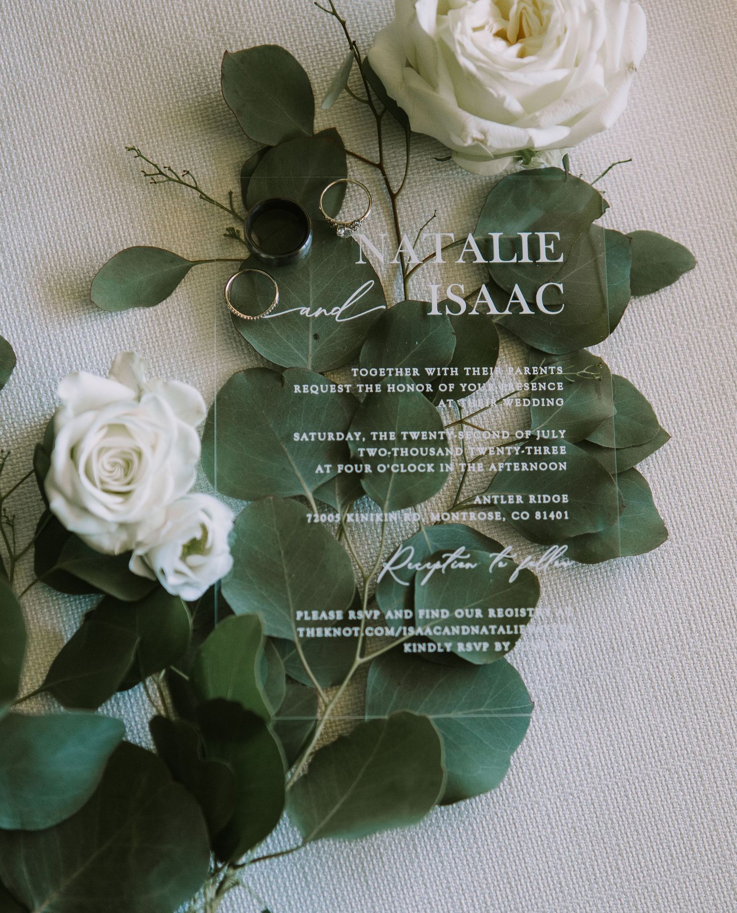 Elegant wedding invitation with white roses and eucalyptus leaves on an acrylic or glass surface.