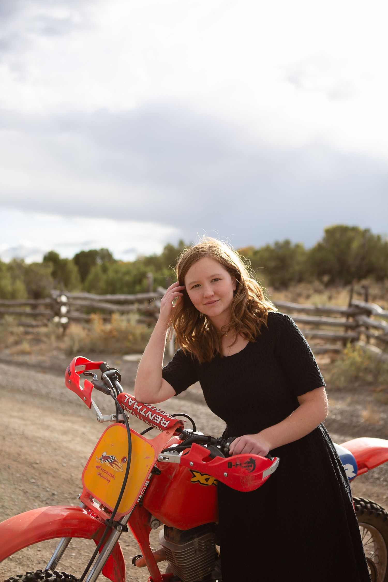 A person in casual wear stands with a red motorcycle against a cloudy rural backdrop.