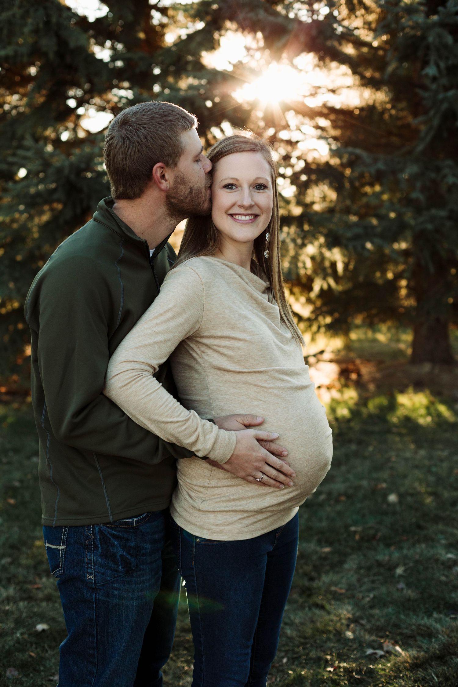 Intimate maternity portrait of couple embracing in front of pine trees at sunset.