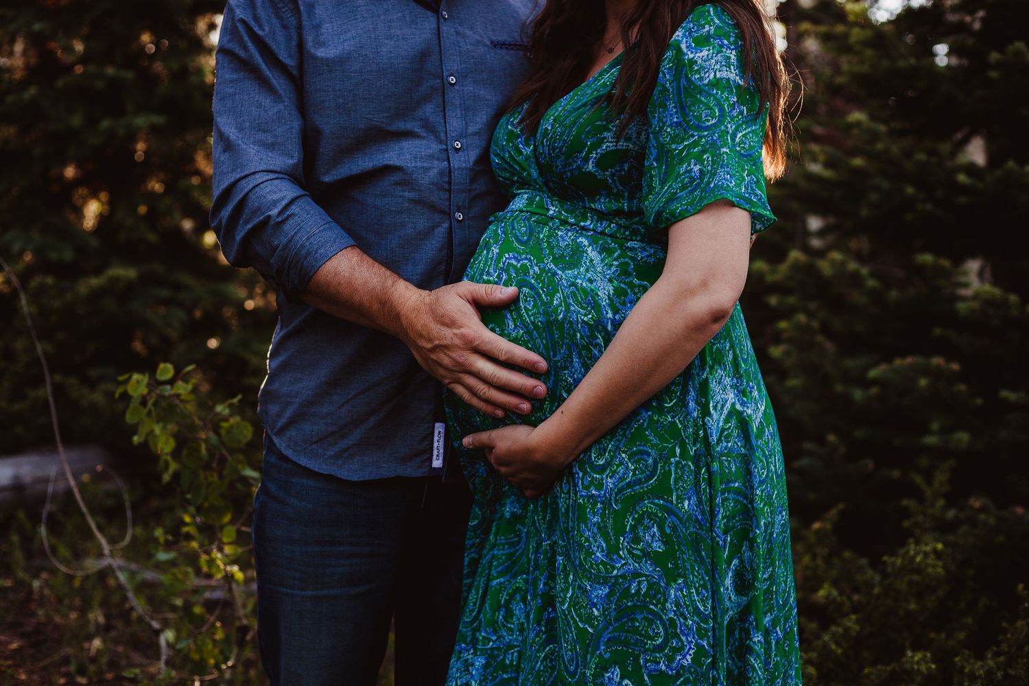 A tender maternity moment captured with hands resting on a pregnant belly in a teal floral dress.