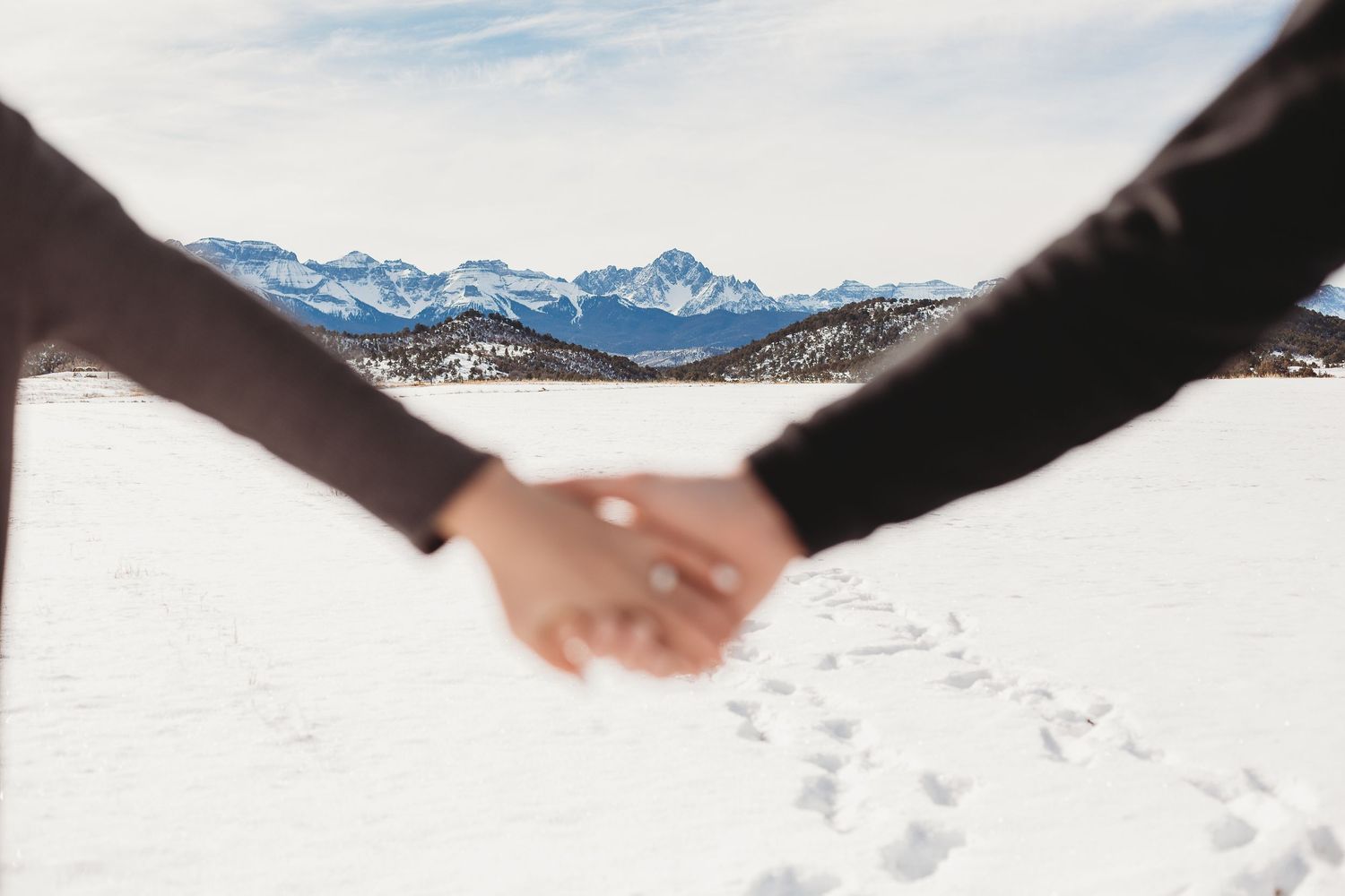 A close-up shot of joined hands with snowy mountain peaks and footprints in the distance.