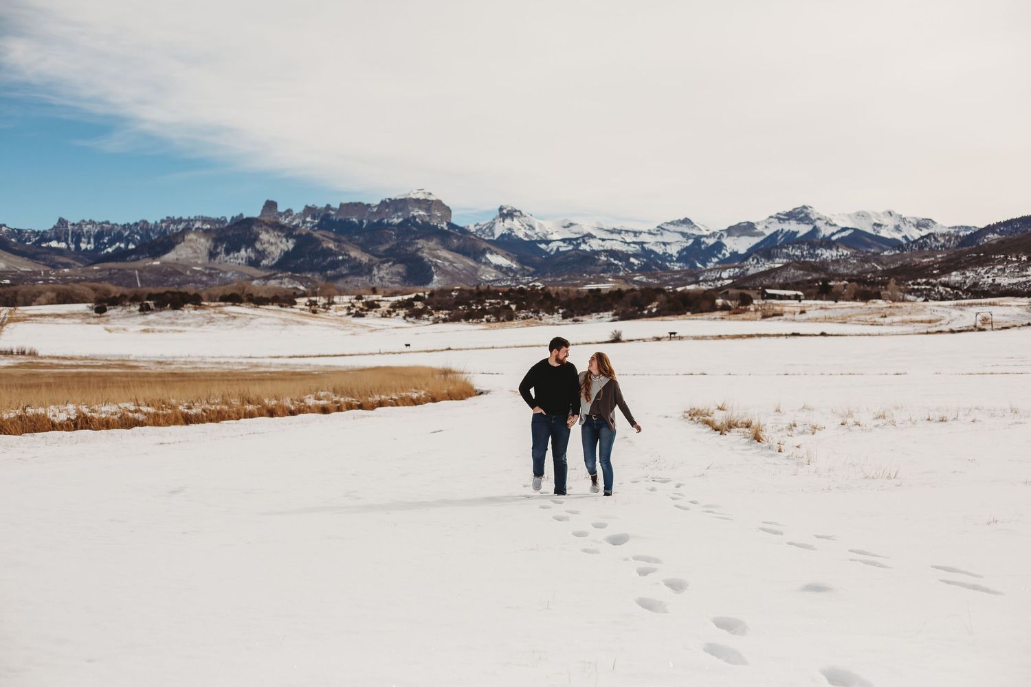 Two people walking together leaving footprints in fresh snow with mountains behind them.