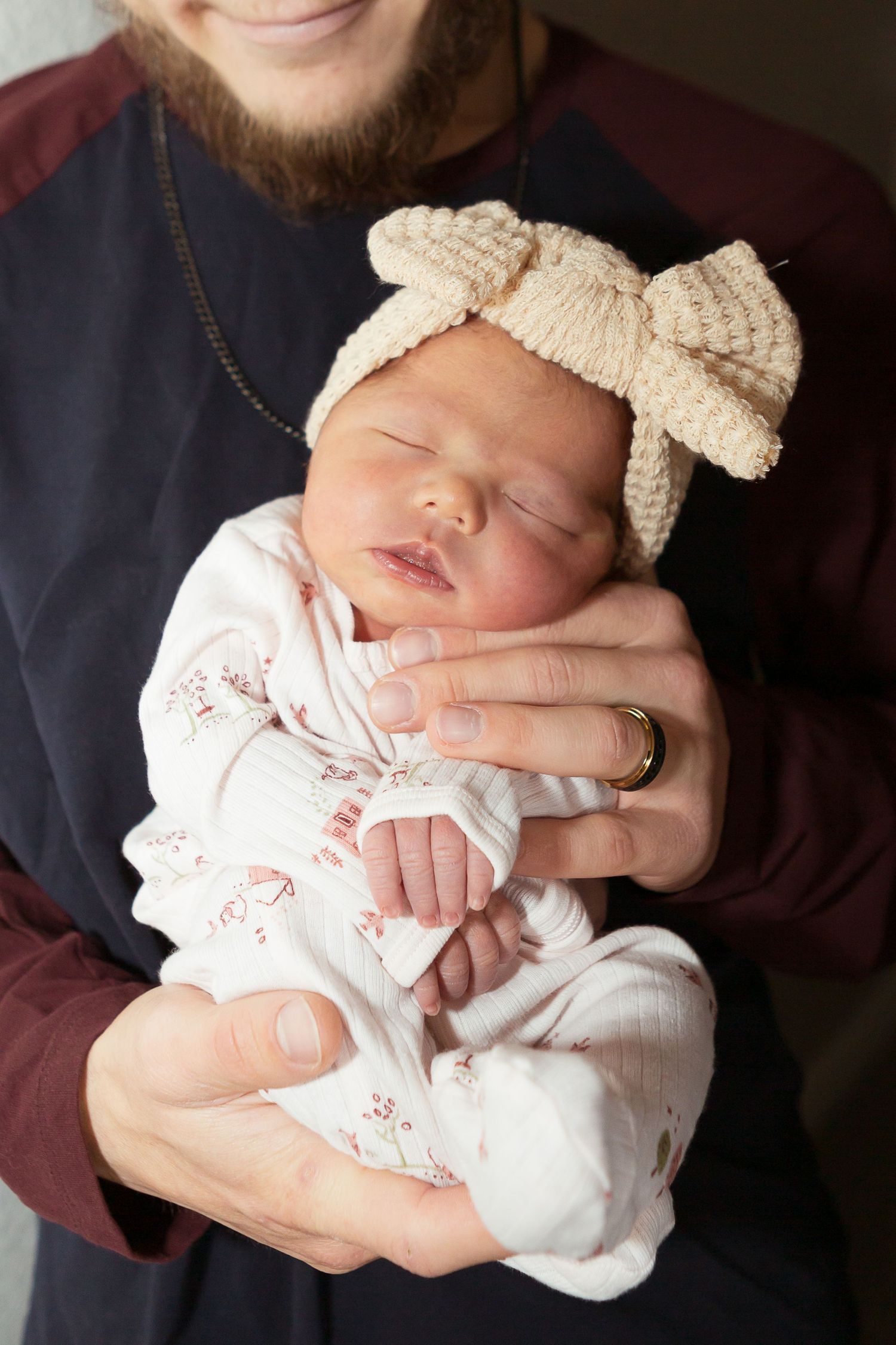 A sleeping newborn baby wearing a cream bow headband and floral outfit being held gently.