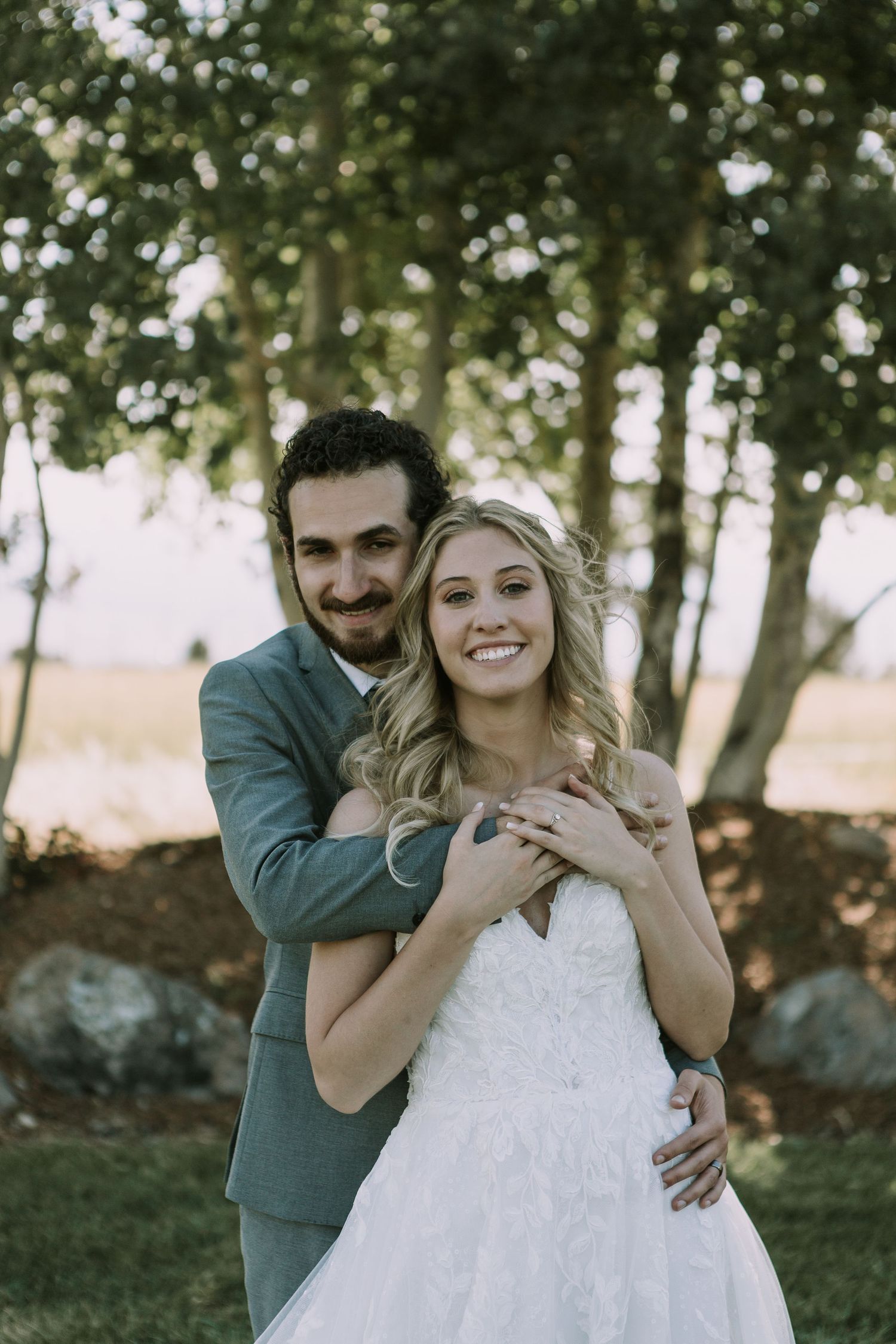 Happy couple embraces outdoors during wedding photoshoot with trees and natural lighting.