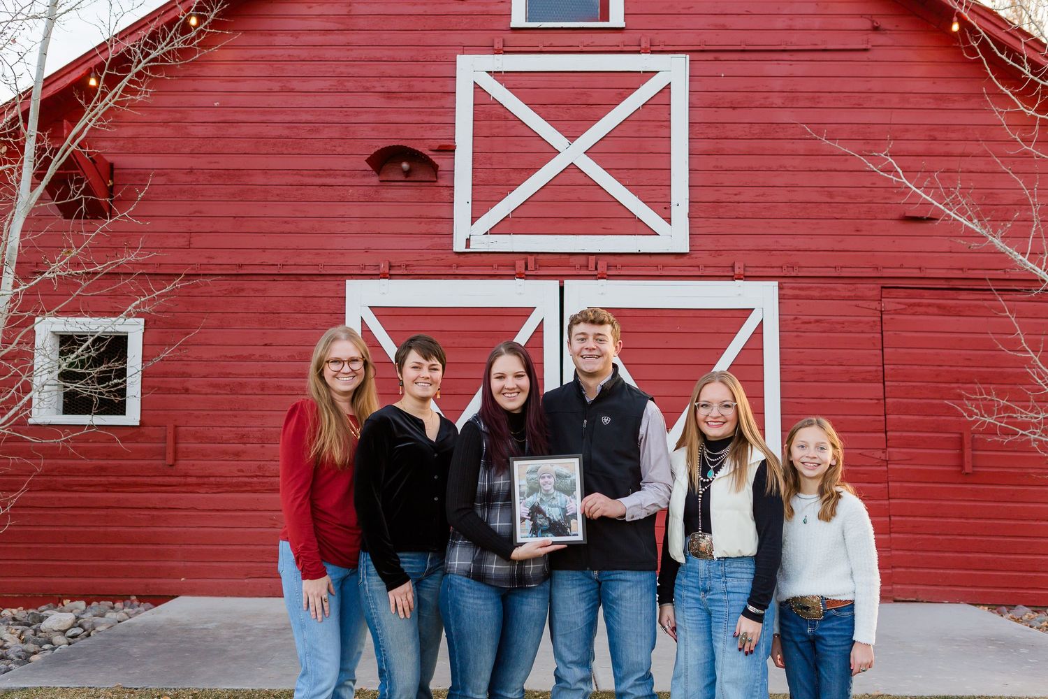 A group shows a framed photo while standing in front of a bright red barn with white X-pattern doors.