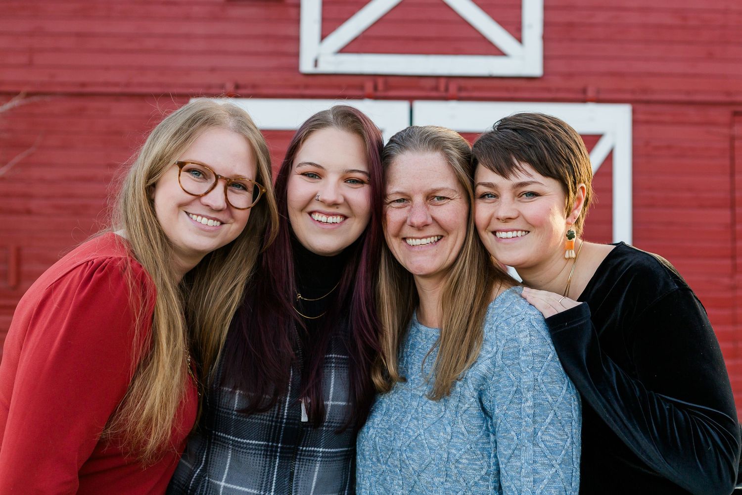 Four friends smile together in front of a red barn with white X-shaped door designs.