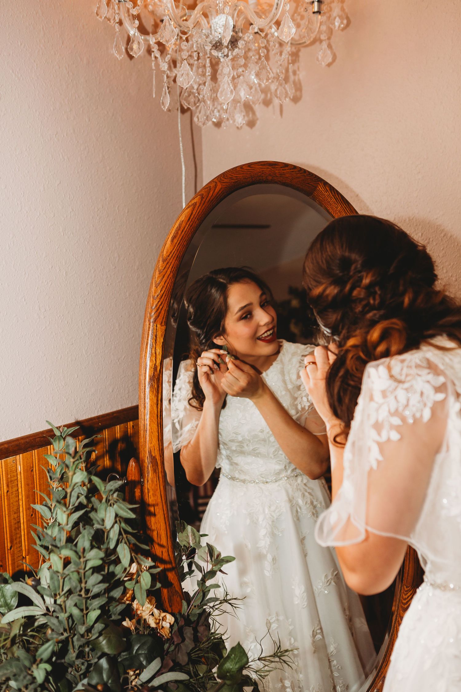 Bride in lace wedding dress puts on earrings while looking in ornate mirror.
