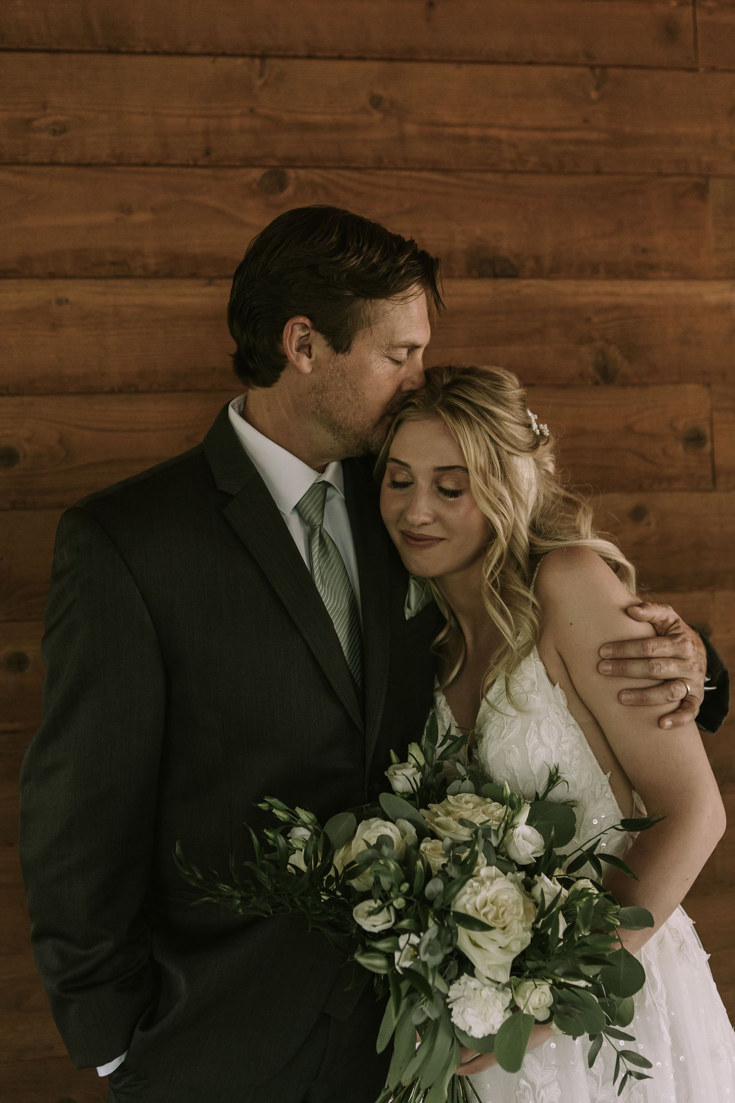 Intimate portrait of a couple in wedding attire posing against wooden wall with white floral bouquet.