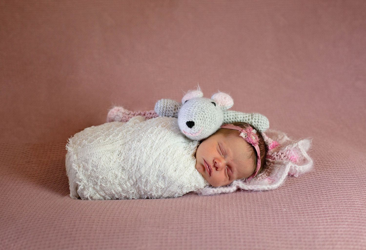 Newborn swaddled in white wrap with small plush toy lying on pink backdrop during professional photoshoot.