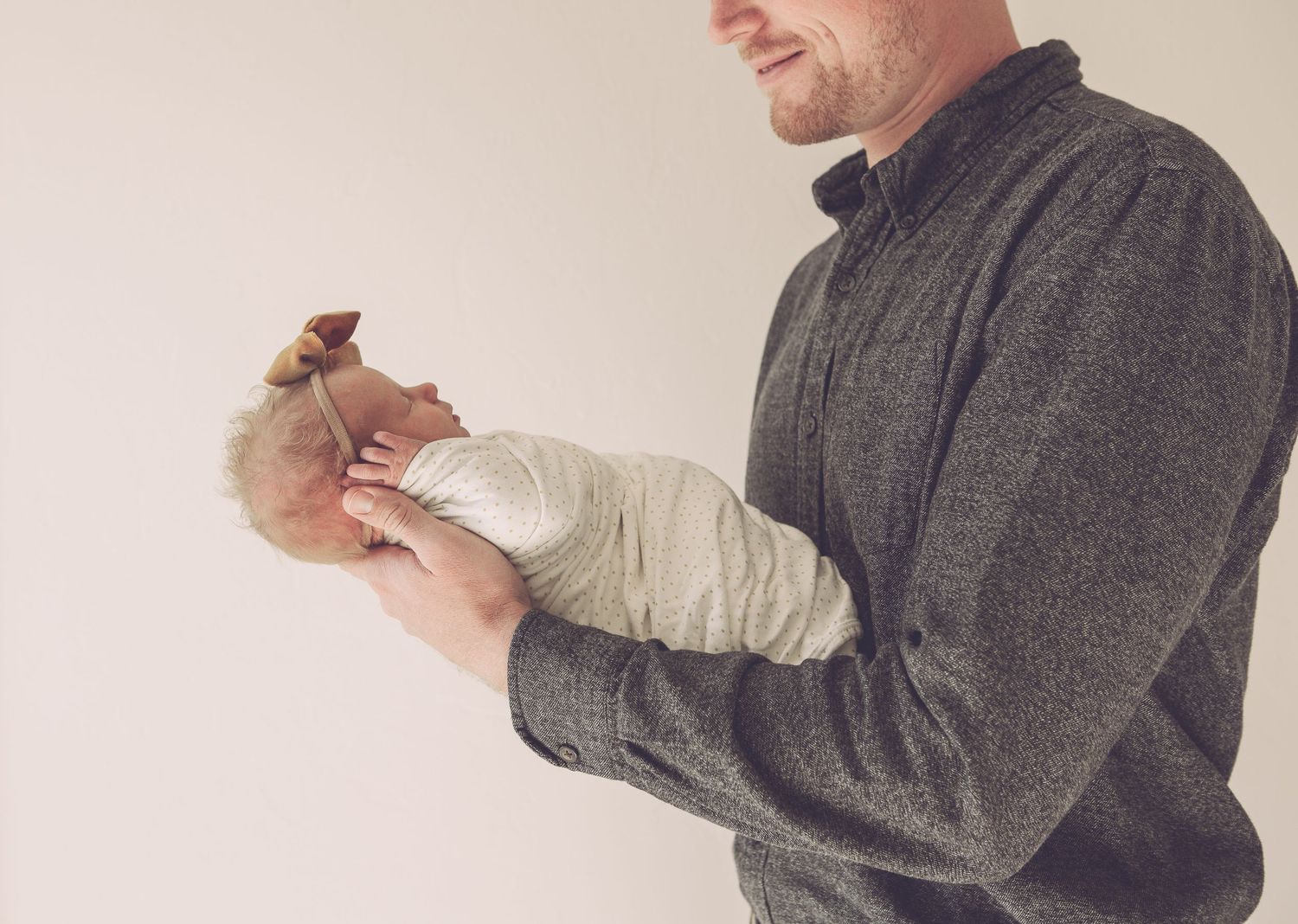 Person wearing grey sweater cradles swaddled infant against beige wall.