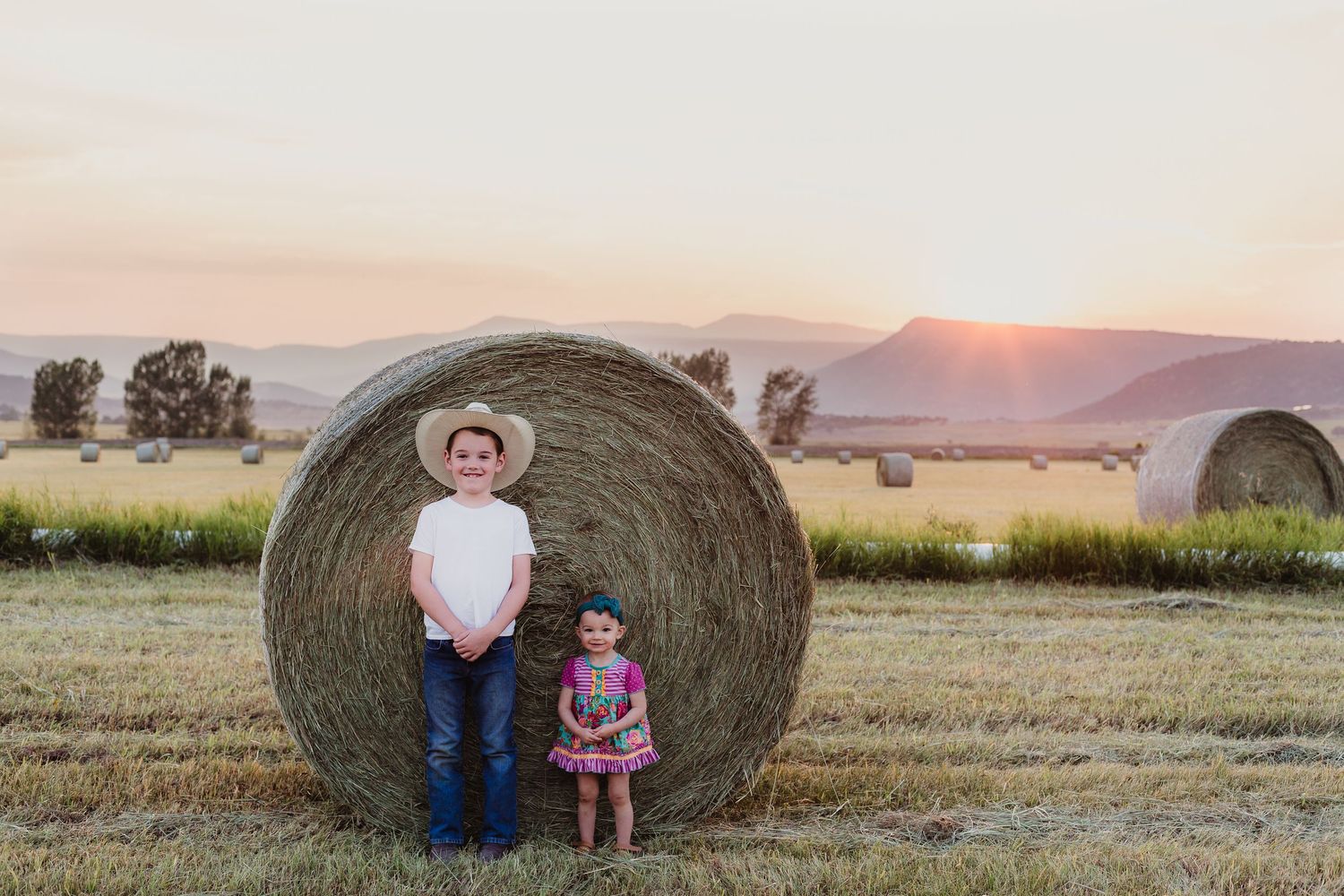 Two people stand against a large hay bale at sunset with mountains in the distance.