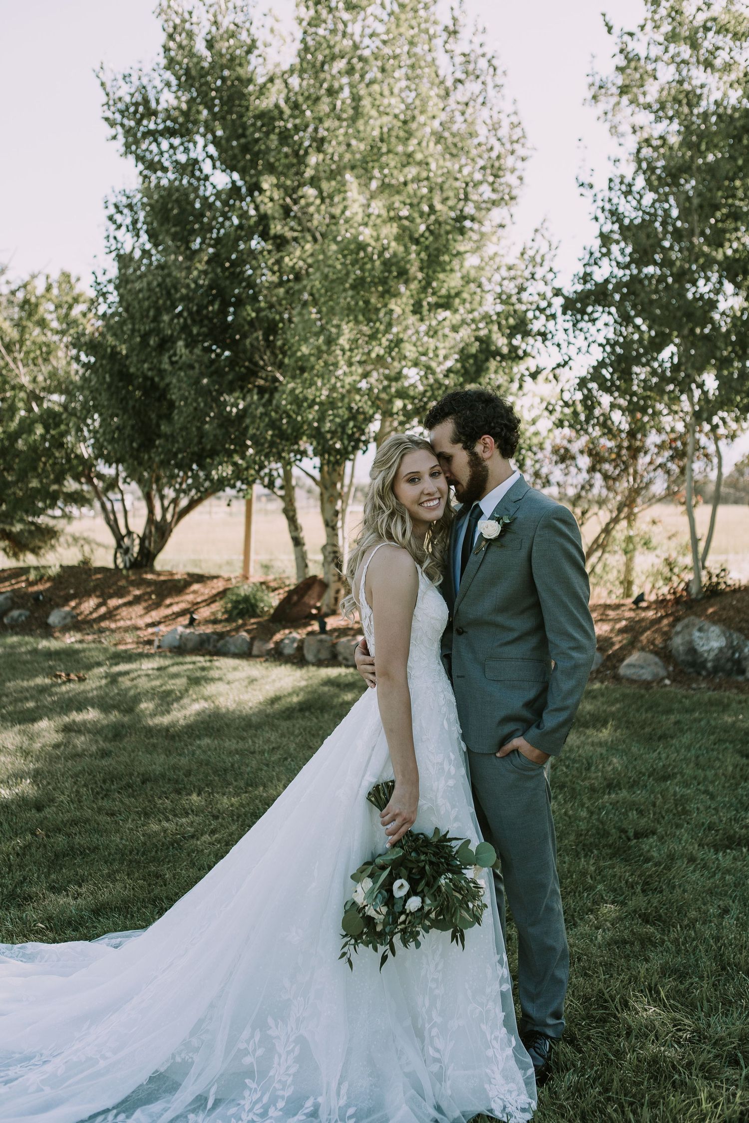 Wedding couple poses with greenery bouquet in outdoor setting with flowing lace dress train.