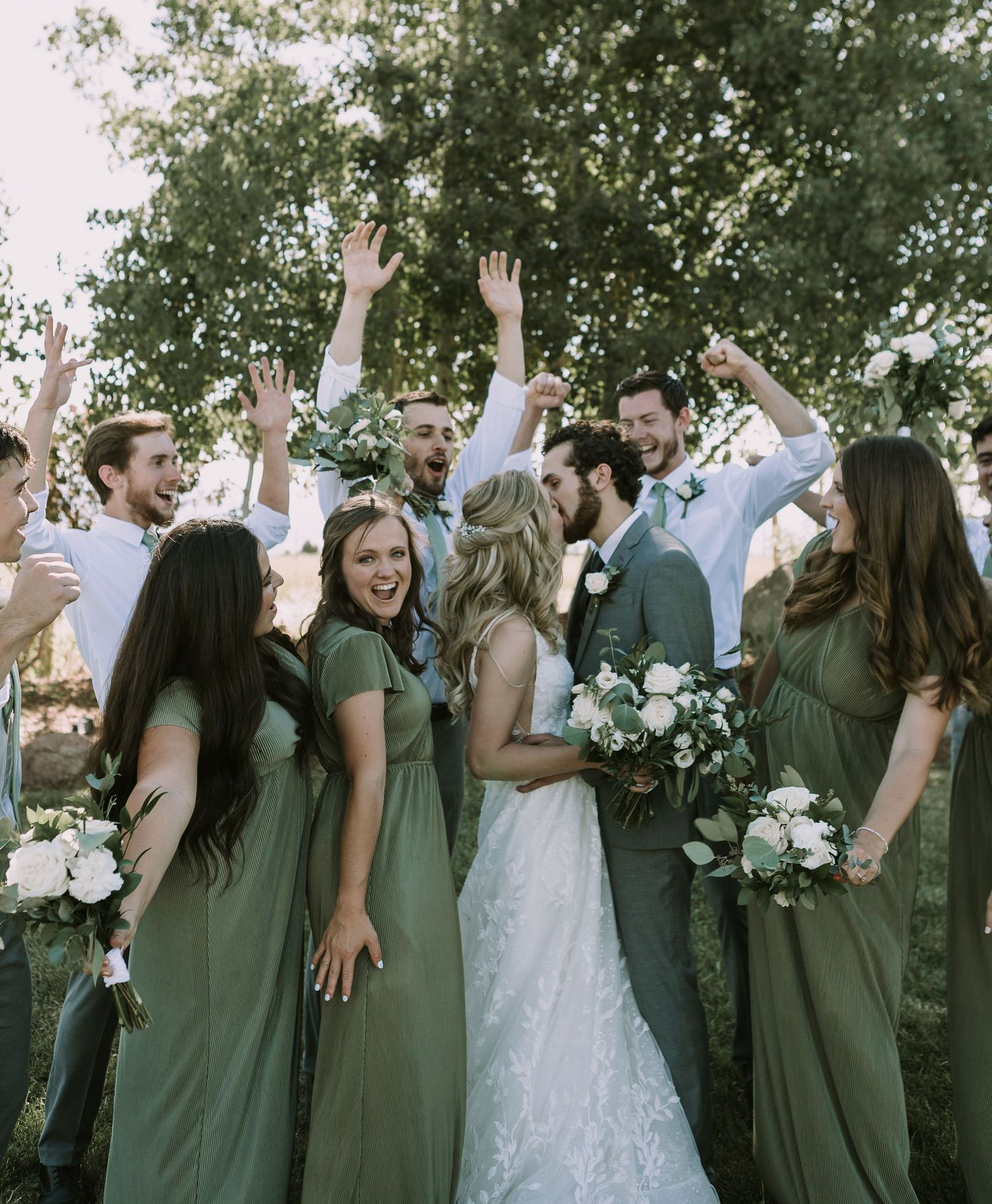 Wedding party celebrates with raised hands outdoors wearing green dresses and gray suits.
