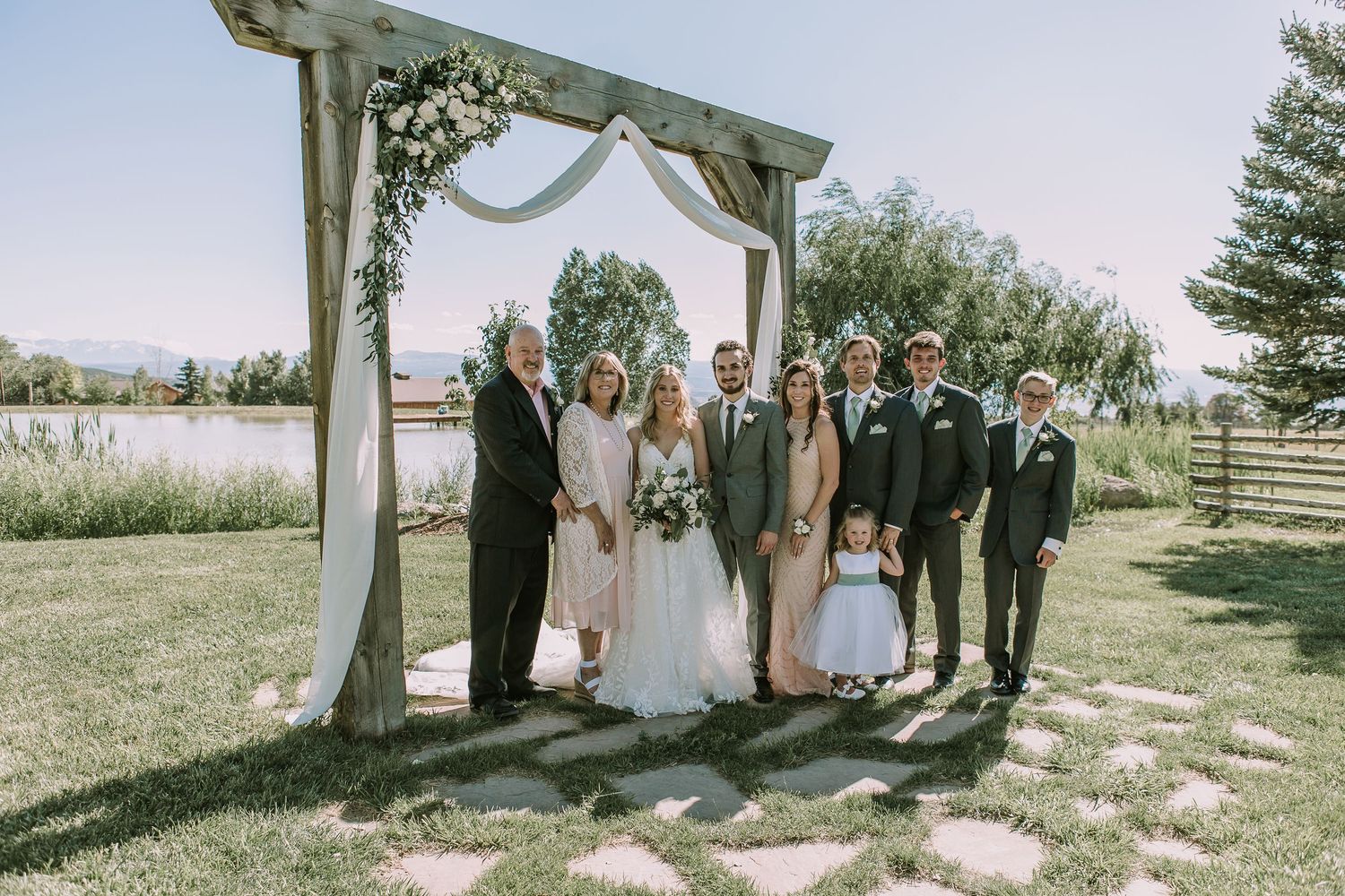 Bridal party gathered under rustic wooden wedding arch on stone pathway in garden setting.