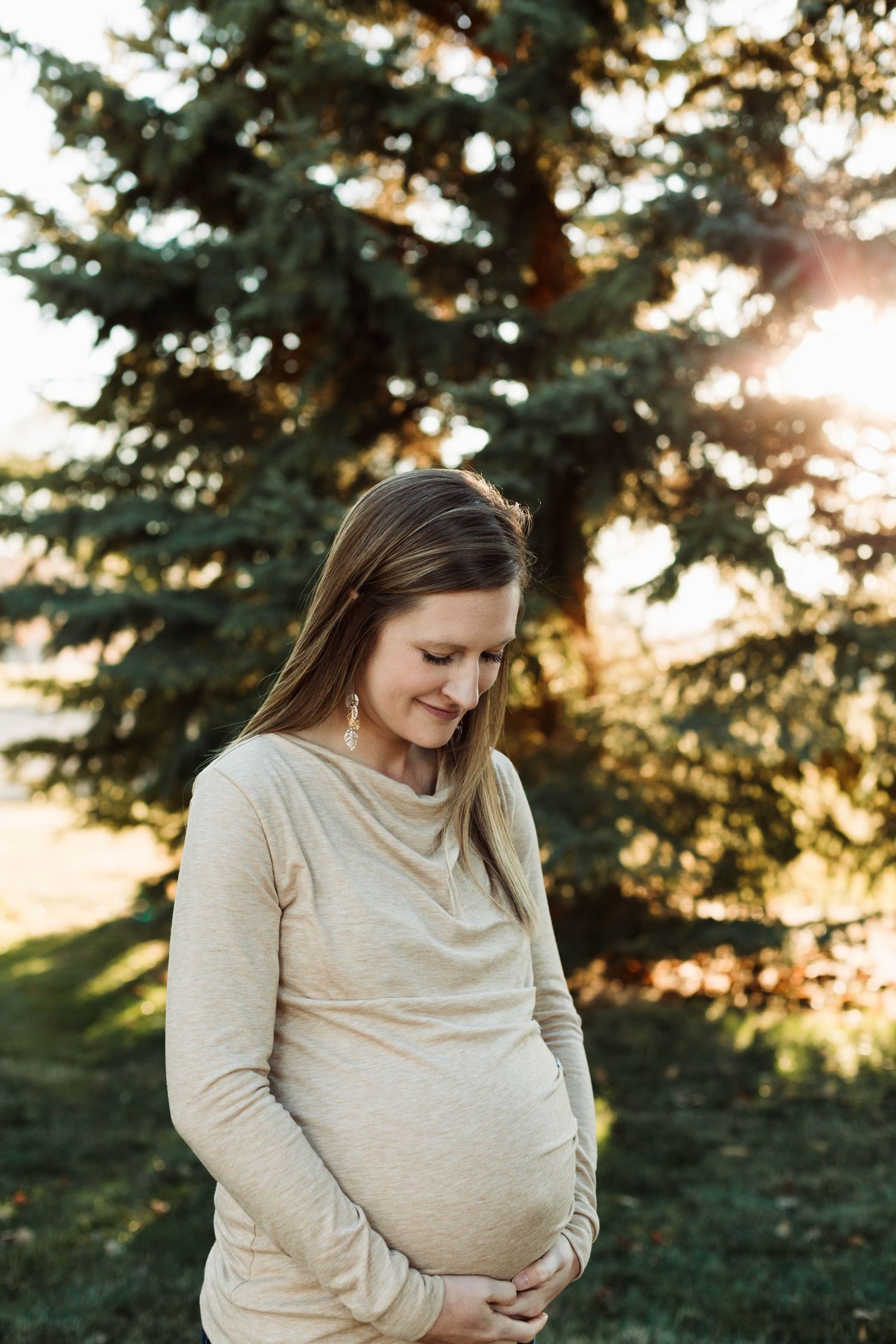 Maternity photo of expectant mother in cream sweater looking down at belly during sunset.