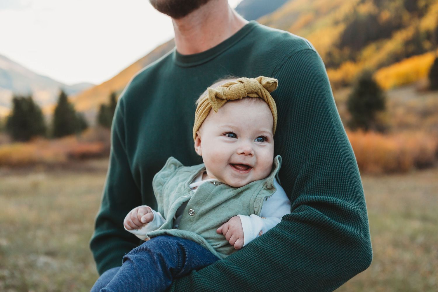 Baby with yellow headband smiling while held in green sweater.