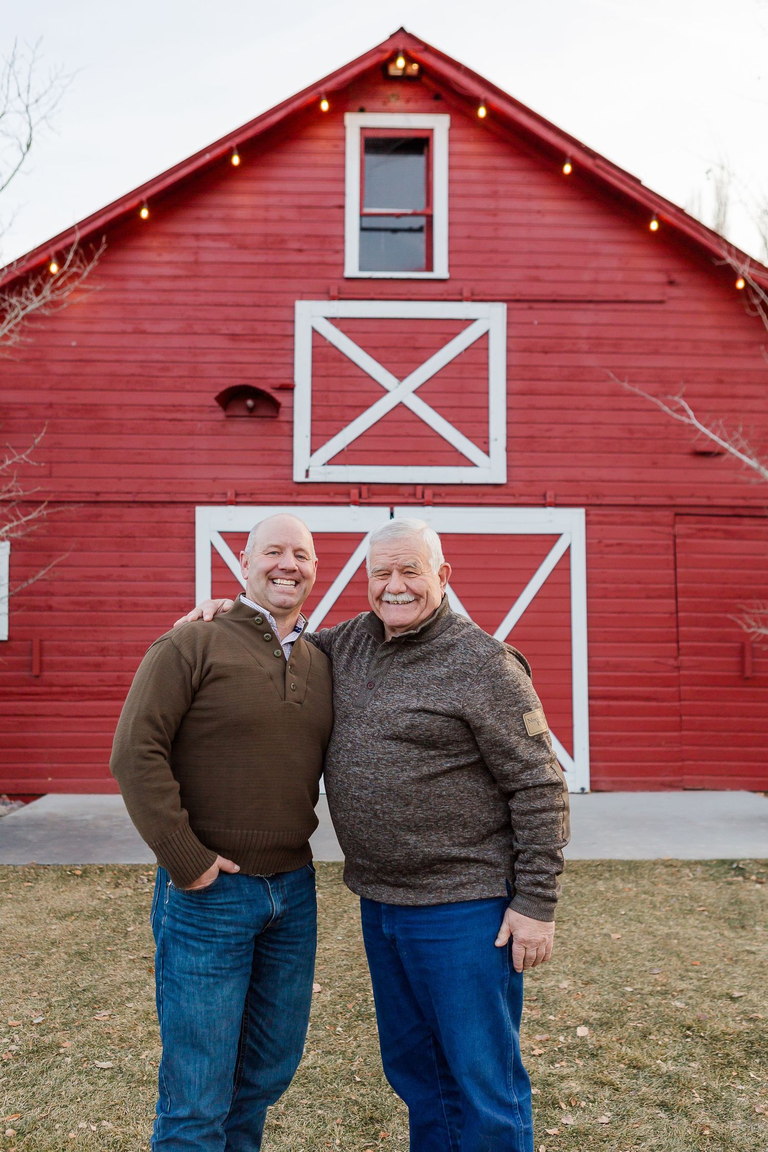 Two people smile together while standing in front of a bright red barn with white trim.