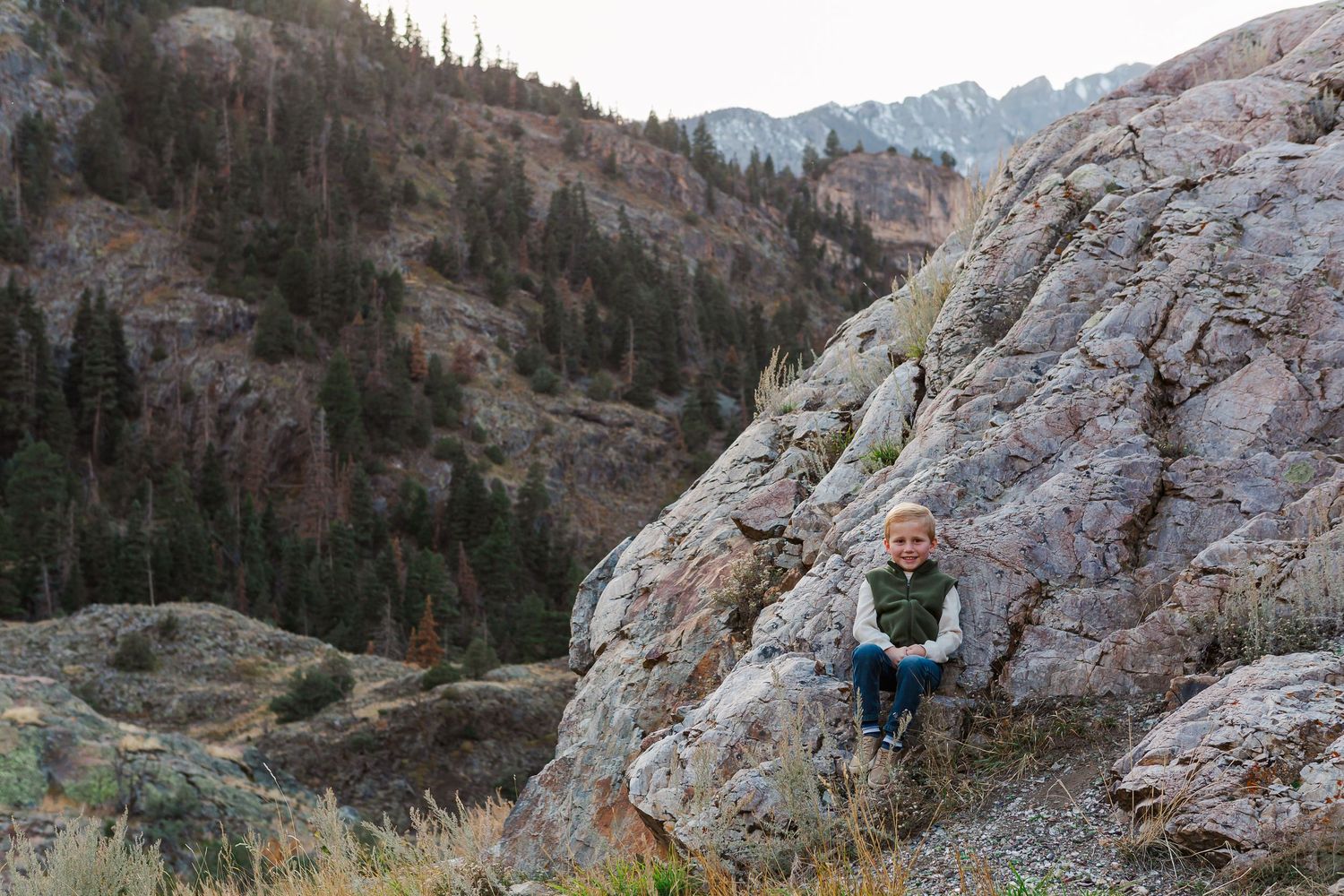 Person sitting on rocky ledge overlooking mountain valley with evergreen trees.
