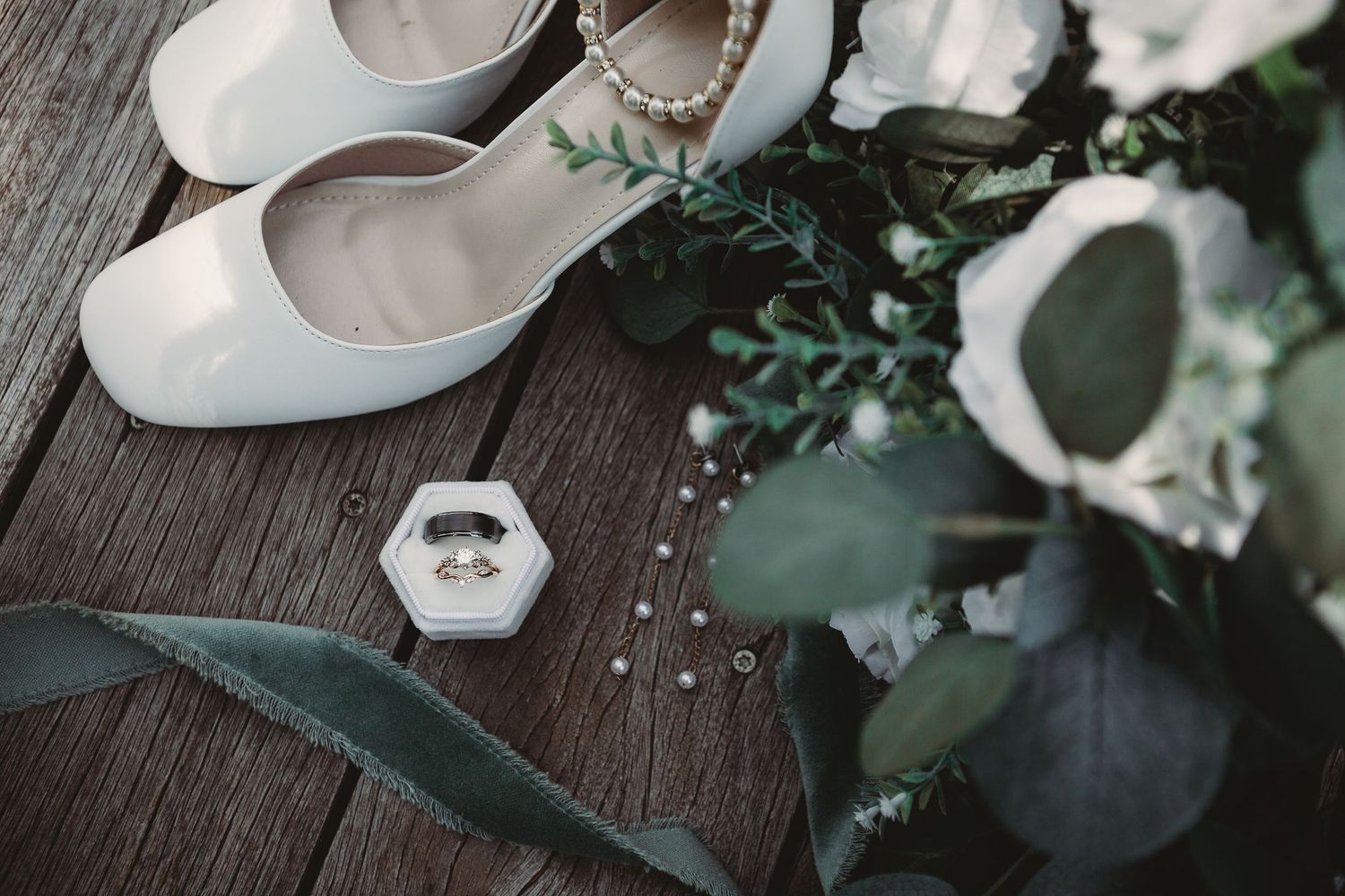 White wedding shoes, a ring box, and delicate floral arrangements on a dark wooden surface.